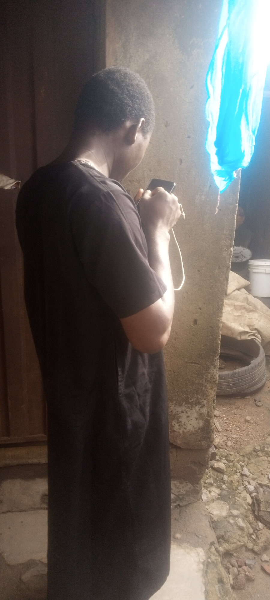 A person stands adjacent to a concrete column, holding a smartphone connected to a white charging cable. The person is wearing a dark, long-sleeved shirt and appears to be looking at the device. A bright blue cloth hangs from the ceiling near the column. A brown door is visible on the left side of the frame. In the background, a tire, a white bucket, and other miscellaneous items are partially obscured by a tan tarp. The ground appears to be littered with debris and stones. The location is presumed to be Yelwa, Nigeria.