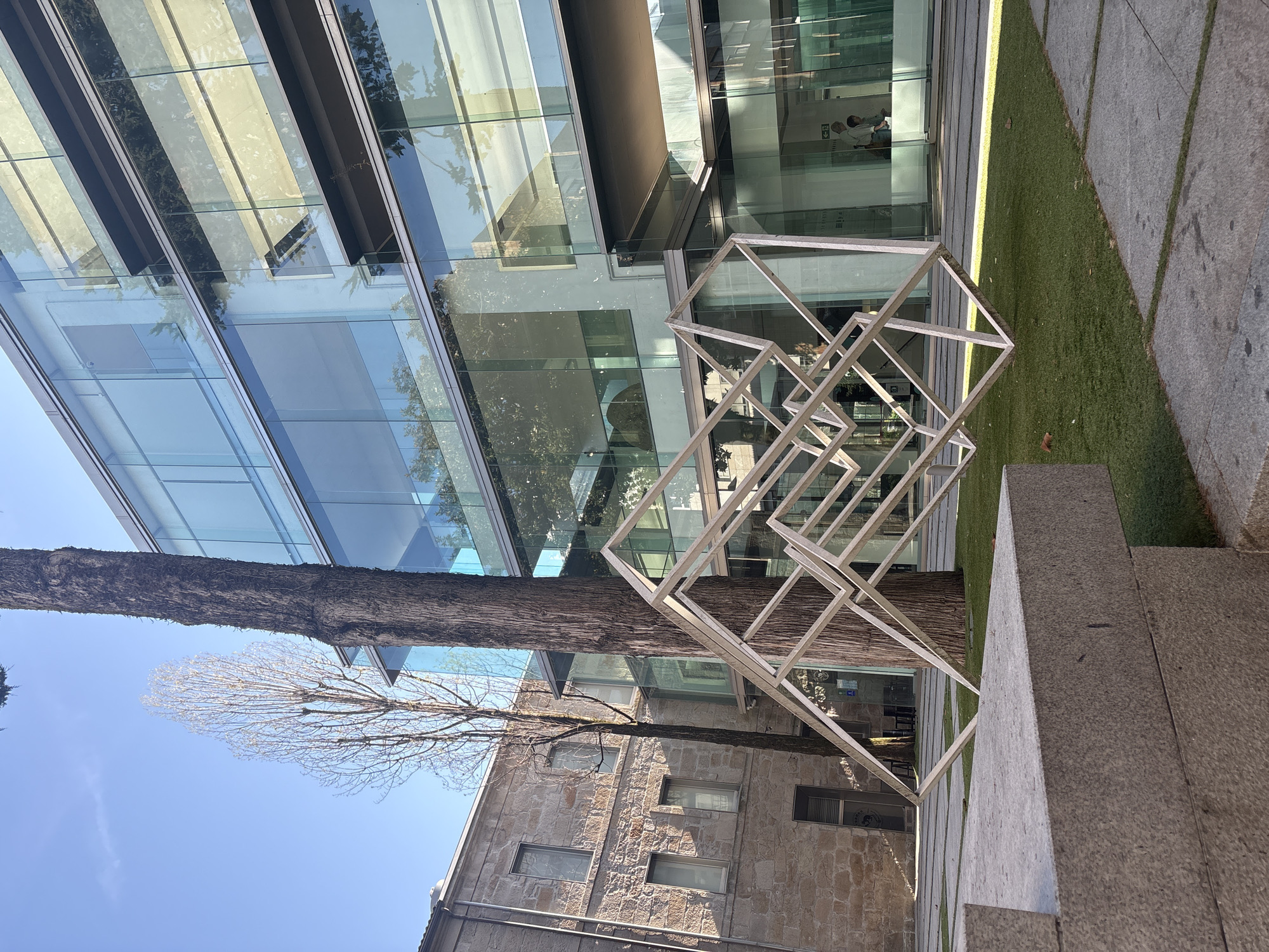 This is a photograph taken in Pontevedra, Spain, in the courtyard of the Cidade da Cultura de Galicia. The image features a modern glass building with reflective windows, a large tree trunk in the foreground, and an abstract metallic sculpture made of interconnected rectangular frames. The sculpture is positioned in front of the tree. To the left of the tree, there is an older stone building with rectangular windows. The ground is a mixture of paved areas and green artificial grass. The sky is clear and blue. There are two people visible inside the modern building through the glass facade.