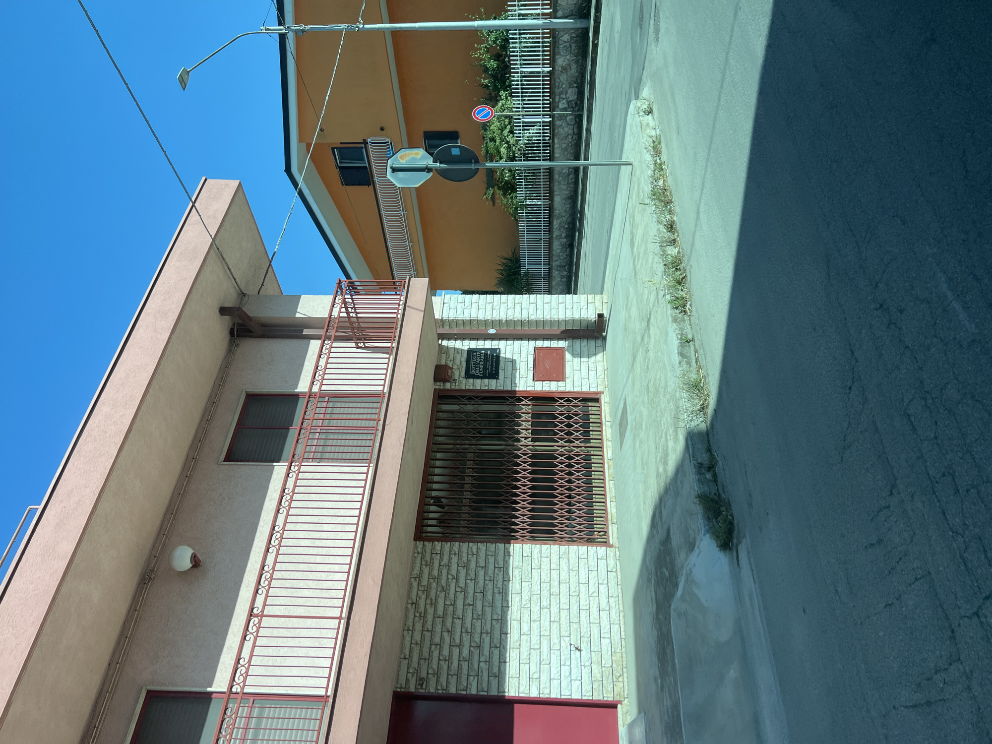 The image depicts a street scene at Via Napoli, 146, in Santeramo in Colle, Italy, under clear blue skies. The primary subject is a two-story building on the left, featuring a light pinkish-beige stucco exterior and a dark red-brown roofline. The second floor includes a balcony with an ornate red metal railing and two windows with dark, possibly shuttered, interiors framed in red. A spherical white light fixture is mounted on the wall above the balcony. Electrical conduits are visible running horizontally across the upper wall.
The ground floor of this building is faced with light-colored, rough-hewn brick. To the left is a red entrance door. To the right of the door, a large window or entrance is secured by a red accordion-style metal security gate. Adjacent to this gated opening, a black plaque with white text is affixed to the brick wall, bearing the inscription "BOTTEGA DELLE FUNE..." (likely "Bottega delle Funerarie" - Funeral Home) and contact information, including a phone number "333.40...". A small white rectangular plaque with the number "146" is also visible to the right of the main sign. Below this, a red rectangular utility box is mounted on the wall.
In the background, to the right, stands a two-story orange building with white eaves and window trim. Several windows are visible, some with white decorative railings or small balconies. A white picket fence runs along the front of this building, partially obscured by green shrubs and small trees.
The foreground shows an asphalt street with visible cracks. A concrete sidewalk and curb run along the front of the pinkish-beige building, with some weeds growing at the edge. A prominent shadow from an unseen object or building covers the lower-left portion of the street, indicating bright sunlight. A metal pole stands near the center-right, supporting multiple traffic signs: an upper, faded octagonal sign, a circular black sign with a white horizontal bar (no entry/forbidden), and a circular red-bordered sign with a blue background and a red diagonal line (no stopping/standing). To the far right, a tall metal streetlight pole with a modern LED fixture is visible, along with overhead power lines traversing the blue sky. No people or vehicles are present in the scene.