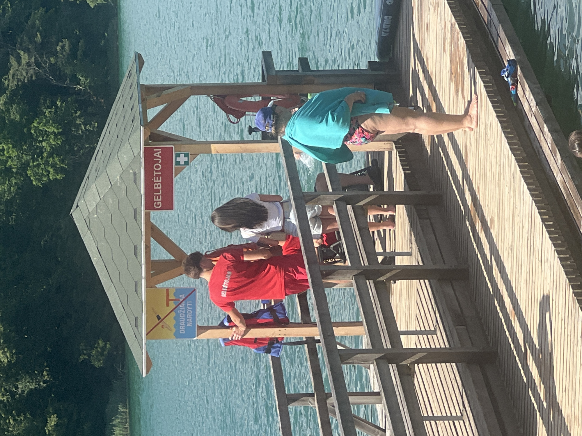 A lifeguard stands on a wooden dock next to a lifebuoy and a life jacket. A woman in a bathing suit and a turquoise towel wrapped around her shoulders, is also on the dock. In the background, a man in a red shirt with "GELBĖTOJAS" printed on the back, stands with his hand on a wooden post. A woman in a white shirt and shorts is standing next to him. A sign above them reads "GELBĖTOJAI" which means lifeguards in Lithuanian. Another sign depicts a person diving and states "DRAUDŽIAMA NARDYTI," meaning diving is forbidden. The scene appears to be at a lake or river in Vilnius, Lithuania, given the signage. The water is a vibrant blue-green.