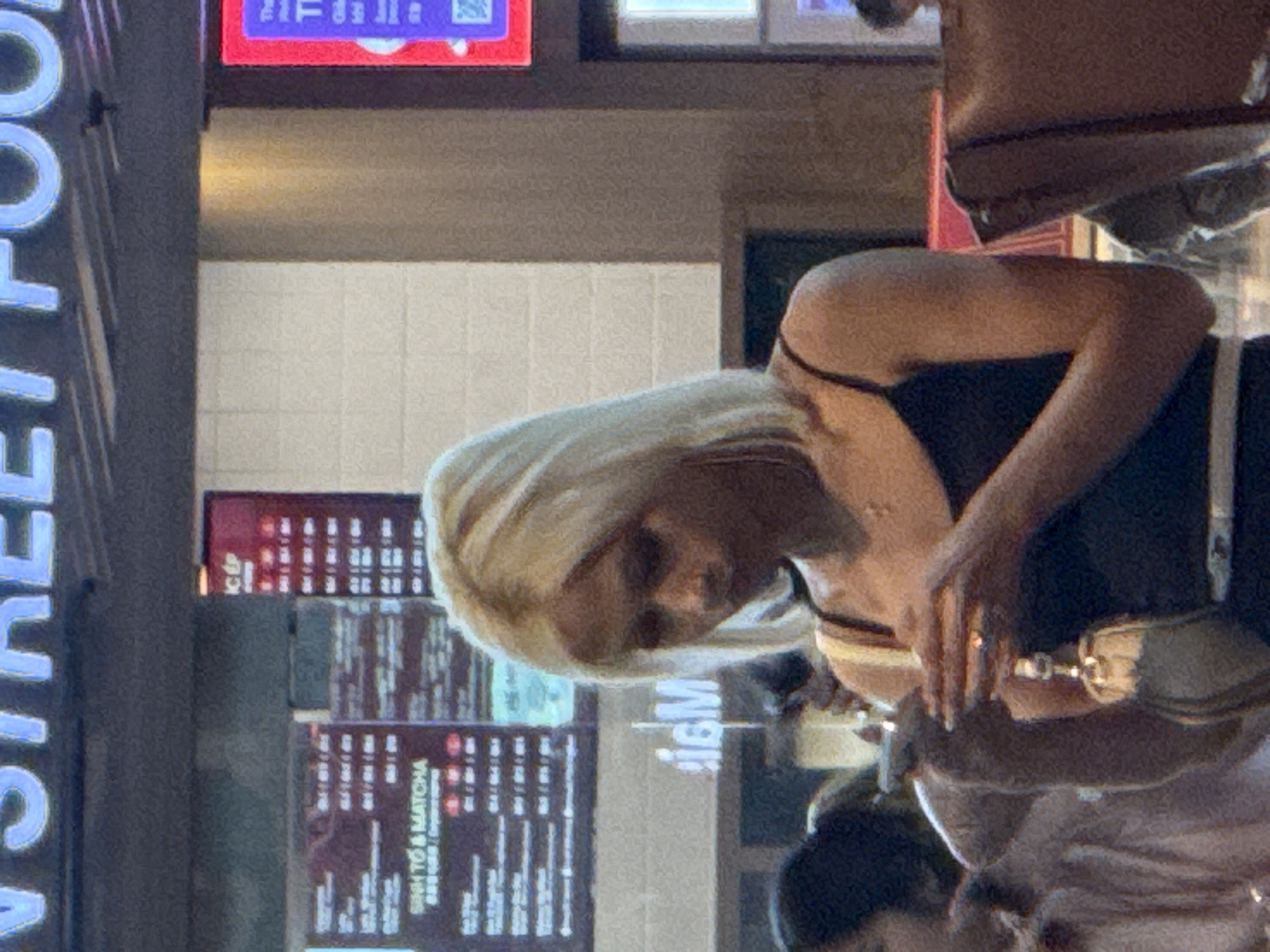 The image shows a young woman with blonde hair, wearing a black tank top, standing in front of a food establishment. She is looking down at a mobile phone she is holding in her left hand, and her right hand is near her face, possibly touching a ring on her finger. A small bag with a tassel hangs from her right arm. In the background, a menu board with Vietnamese text is visible, listing items like "SINH TO & MATCHA". Above the menu, a sign with large blue letters reads "STREET FOOD". The environment appears to be an indoor area, possibly a food court or a restaurant, with tiled walls and signage. The lighting is dim, with some bright lights from the signs and screens. The presence of Vietnamese text on the menu board strongly suggests the location is within Vietnam. Given the context clues such as the signage and the food offering, the image is likely taken at a food establishment in the city of Bắc Cam Ranh, Vietnam.