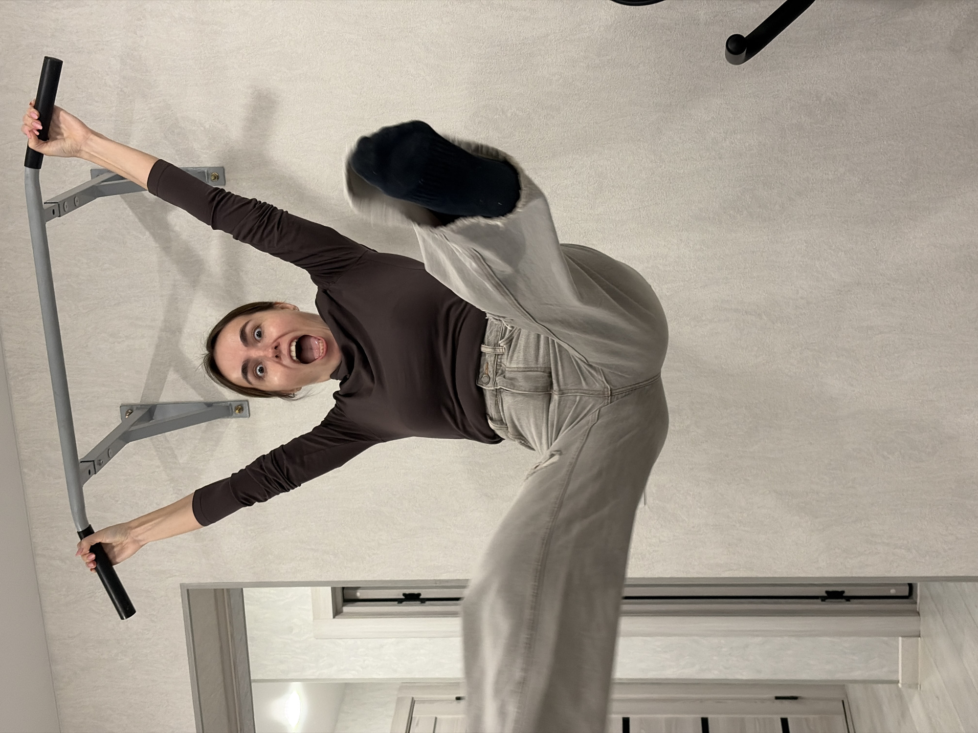 A woman is hanging upside down from a pull-up bar mounted to a wall. She is wearing a brown long-sleeved shirt and light gray pants. She has one leg extended, with a black sock visible. Her mouth is open in an exaggerated expression. The background includes the wall to which the pull-up bar is attached, part of a doorway, and the top of a chair. There is no evidence to suggest that the image was taken in the city of Sarapul, Russia.