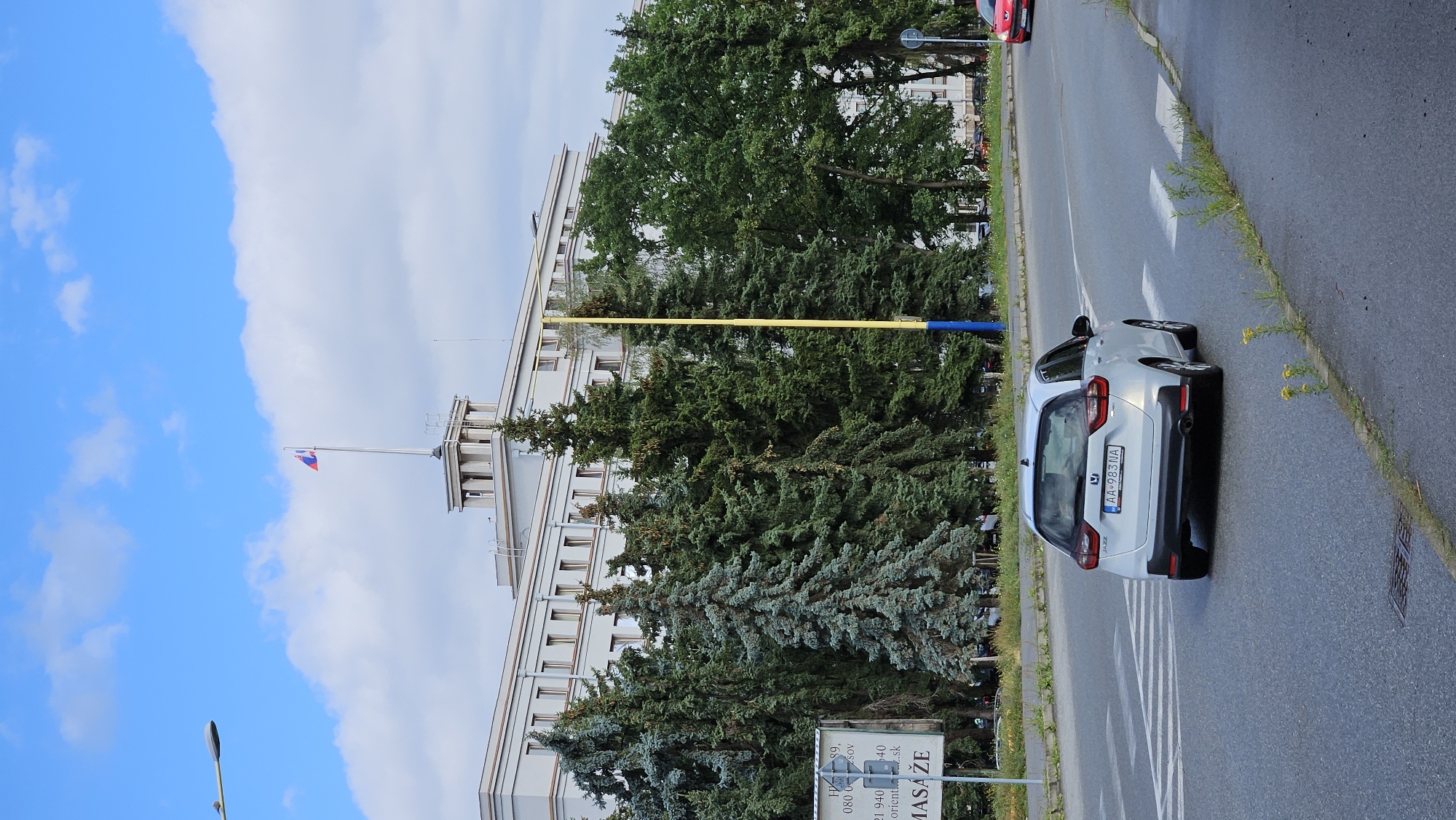 A white Honda Jazz car is driving on a road in front of a large, light-colored, modernist building. The building has many windows and is topped by a tower with a flag on top. Several tall evergreen trees are planted in front of the building. A yellow pole with a blue base is also visible near the trees. On the left side of the road, there is a sign with some text and numbers on it, including "MASÁŽE". The road has white pedestrian crossing markings. A red car is visible in the distance on the right side of the road. The sky is blue with some white clouds. The image appears to be taken in the city of Prešov, Slovakia, likely on a street leading to or from a government or institutional building.