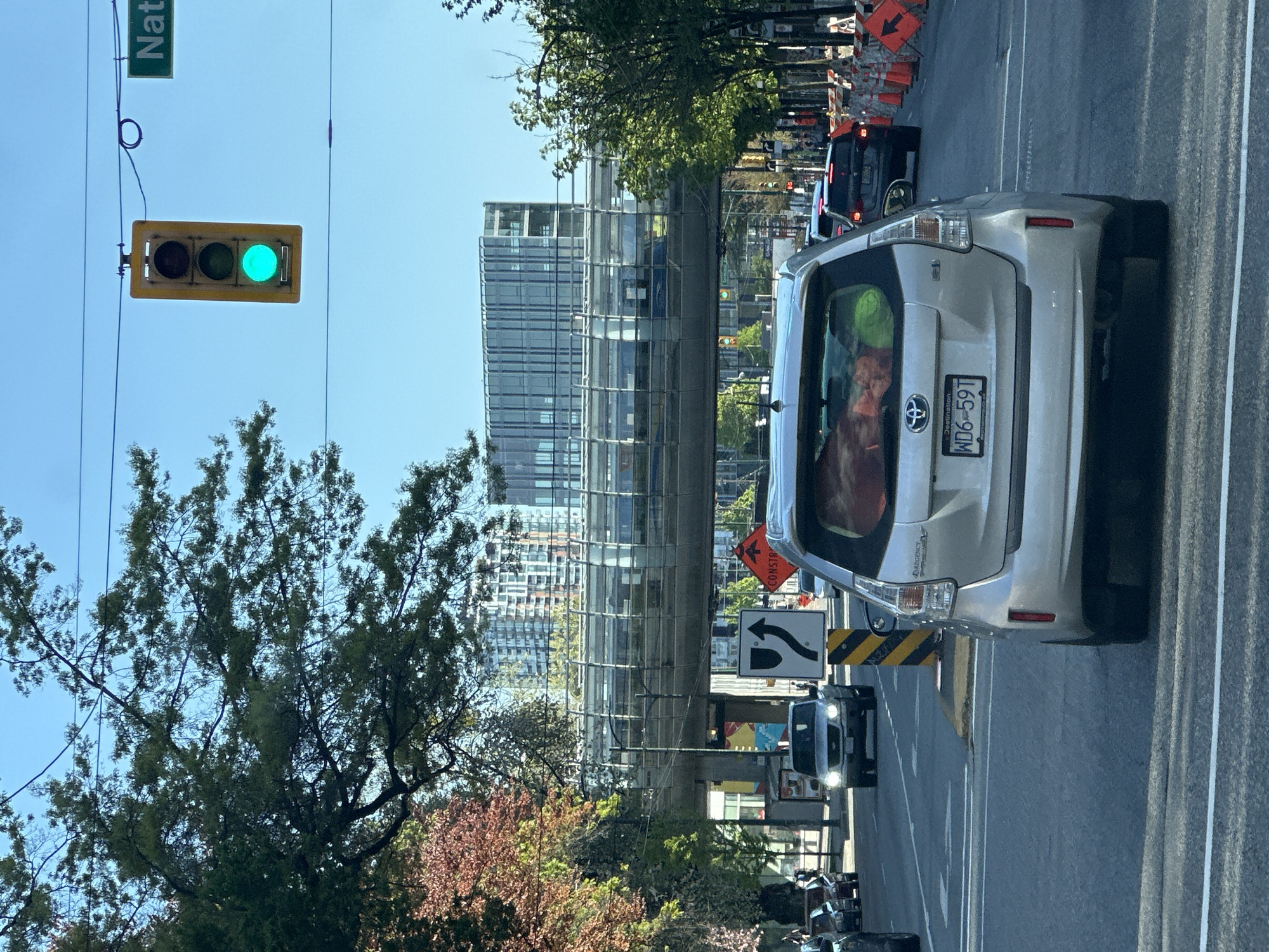 This image shows a street scene in Vancouver, Canada, likely taken from the perspective of someone in a car stopped at a traffic light. The traffic light is green. The street appears to be congested, with several cars ahead and behind the vehicle from which the photo was taken. In the foreground, there is a silver Toyota Prius with the license plate "WD6-59T". The rear of the vehicle is facing the camera, revealing the cargo area filled with items. A modern glass-covered building can be seen in the background, likely part of the Canada Line infrastructure. There is a street sign in the upper left corner that reads "Nat". Construction signs and cones are visible on the right side of the road, suggesting ongoing road work. A tree with green and reddish leaves occupies the left side of the frame. The sky is clear and blue.