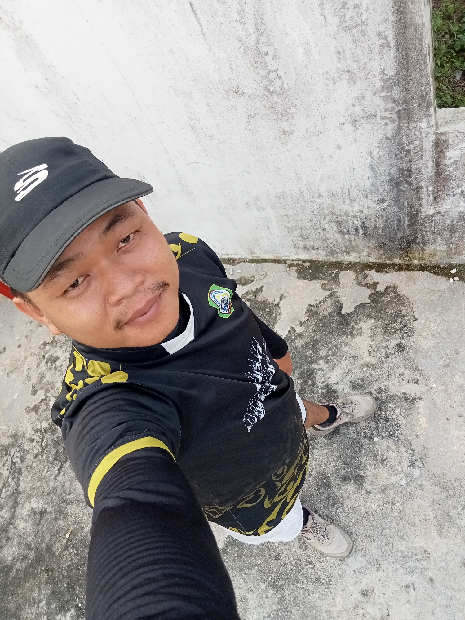 In Kualu, Indonesia, a self-portrait shows a person from a high angle, wearing a black baseball cap with a white logo, a black long-sleeved shirt with yellow accents and a crest on the left chest, and tan athletic shoes. The background consists of a worn, textured concrete surface and a light-colored, distressed wall. The person is standing on the concrete ground, with plants visible in the upper right corner behind the wall.