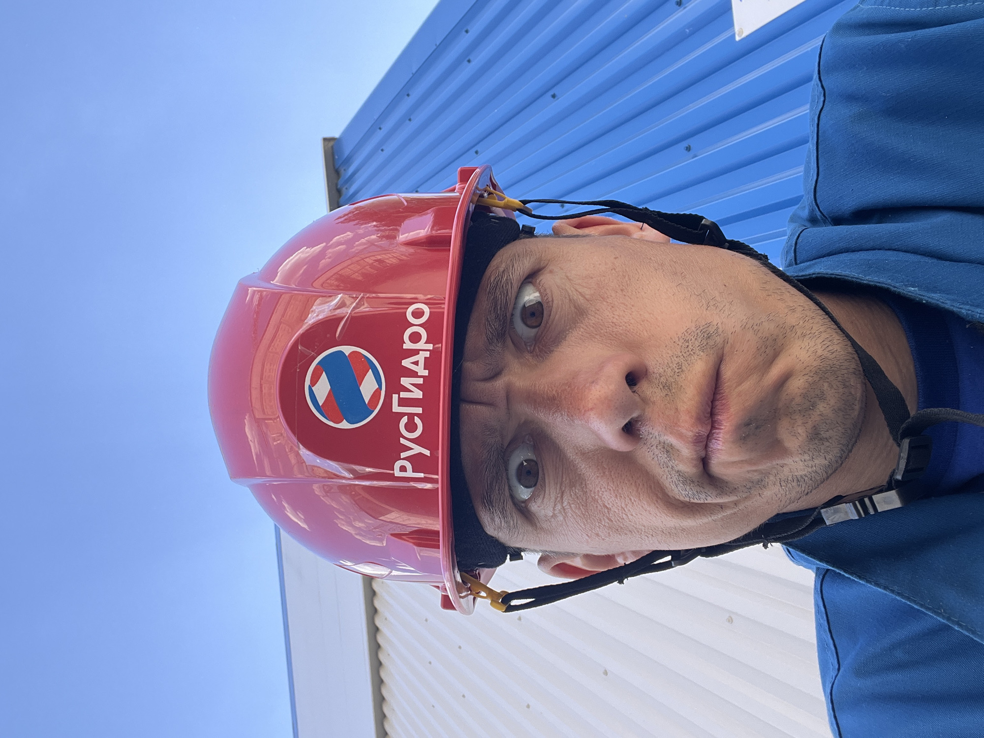 A close-up, slightly rotated selfie shows a man wearing a red hard hat with the logo of "РусГидро" (RusHydro) on it. He has short dark hair, light brown eyes, and is wearing a blue work shirt. The background consists of a blue corrugated metal wall with white trim and a clear blue sky. The location is the city of Chaikovsky, Russia.