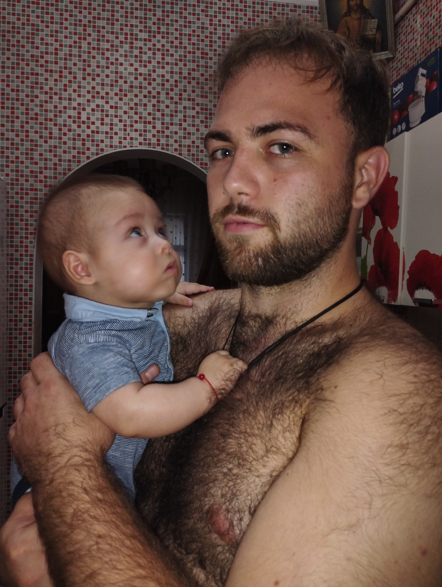 A shirtless man with dark, short hair and a beard holds a baby. The man is facing the camera, and the baby is looking up and to the left. The man's chest and arms are covered in dark hair. The baby is wearing a light blue striped shirt. Behind them is a tiled wall with red and grey squares, and a framed picture on the wall. A box labeled "Beko" is visible on the right. The overall impression is an indoor setting, likely a home. The image does not contain information indicating its location in Fălești, Moldova.