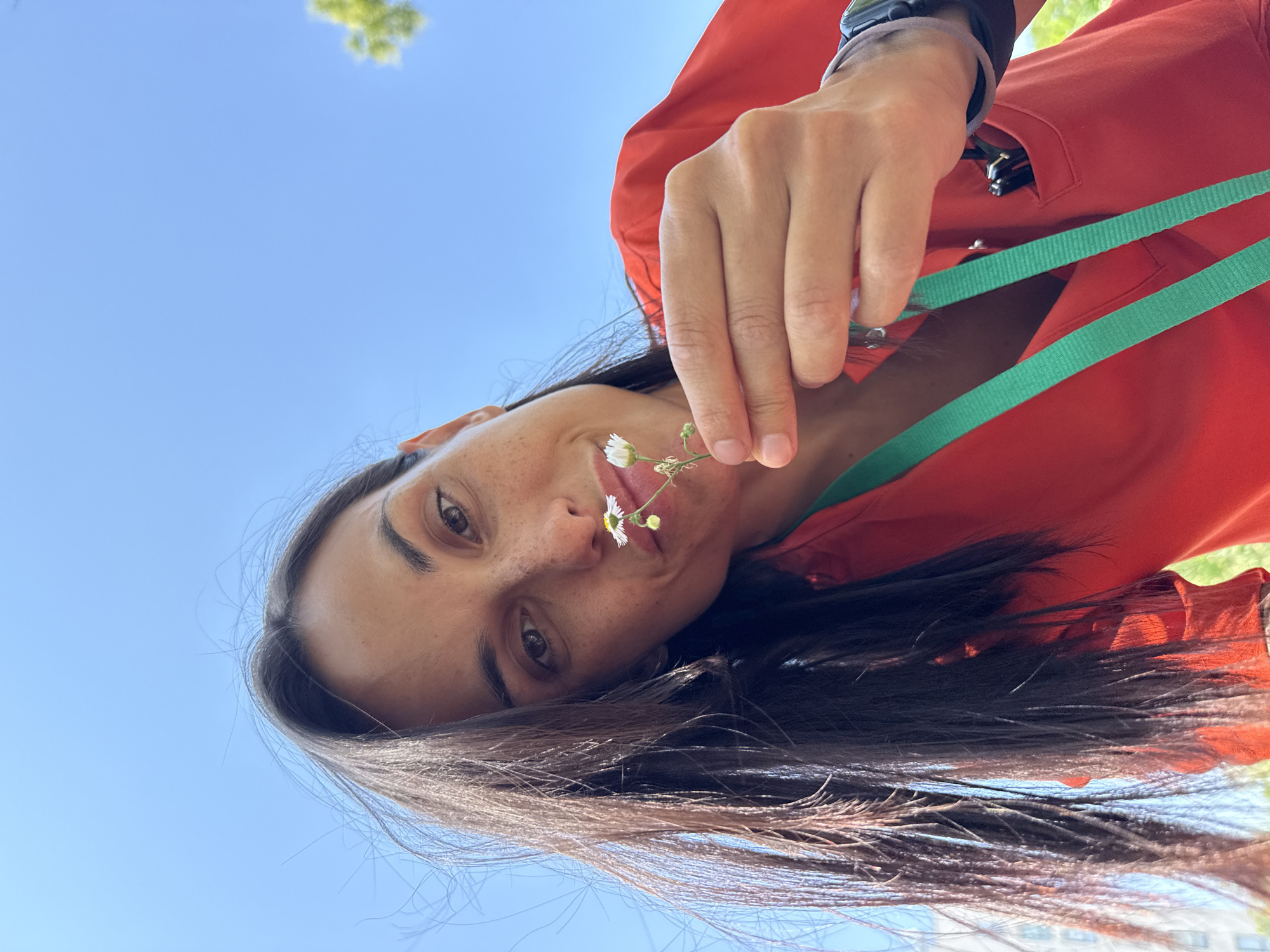 A woman, appearing to be in her late 20s or early 30s, is captured in a low-angle, selfie-style shot against a bright blue sky. She has long, dark hair and is wearing a vibrant red garment, possibly a uniform or scrub top, with a green lanyard around her neck. She holds a delicate white wildflower up to her nose, her lips slightly pursed. The woman has a smattering of freckles across her nose and cheeks. Her right hand is visible, holding the flower stem. A black watch is worn on her left wrist. In the background, a blurry green tree branch is visible on the right, and to the left, a building with visible window frames suggests an urban or semi-urban setting. The overall impression is one of a casual, outdoor moment, possibly during a break or a walk, with the city of Pleven, Bulgaria, being the implied location due to the context provided.