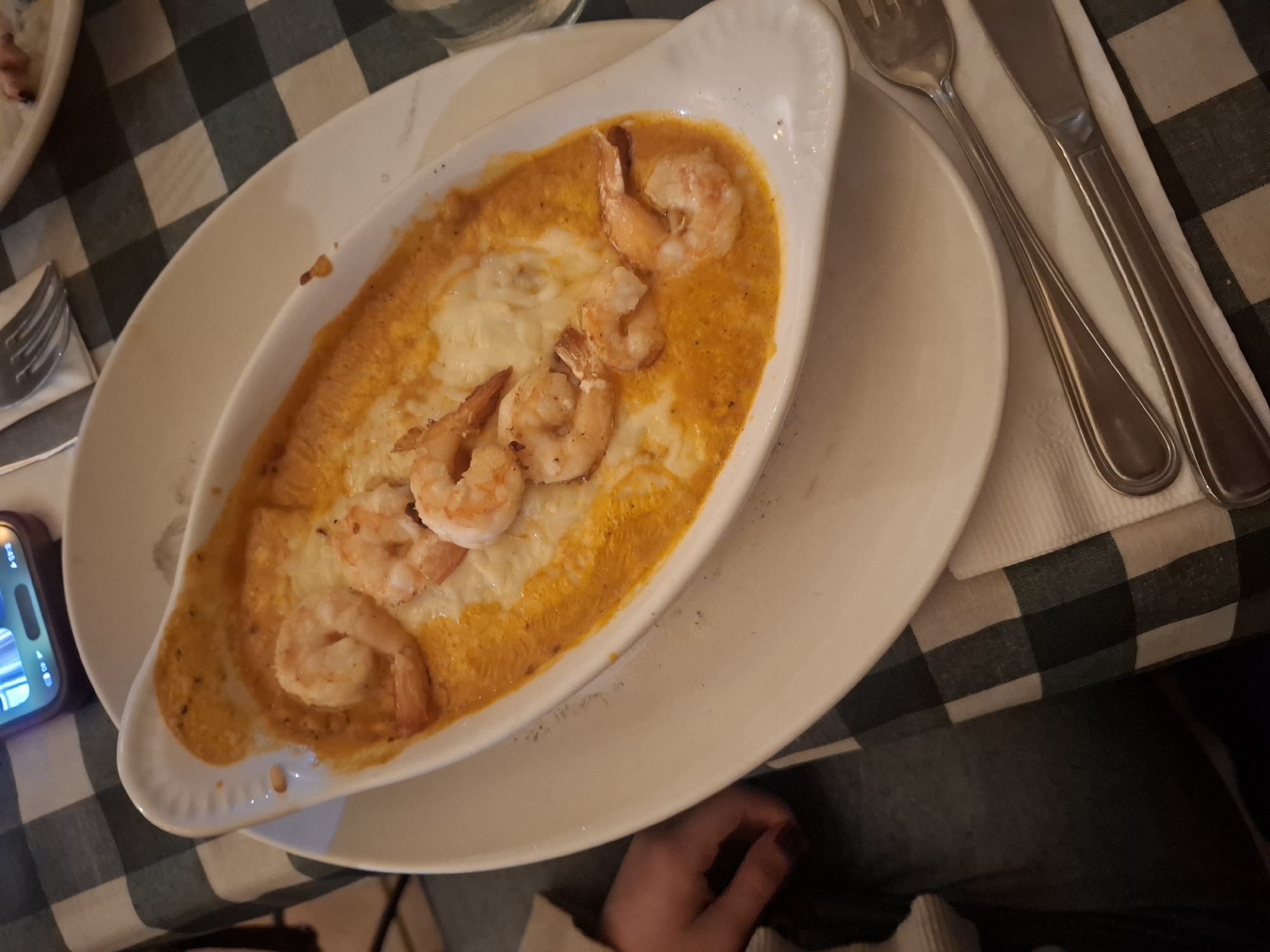 A top-down shot shows a plate of shrimp and grits in a white oblong dish on top of a larger white plate. A fork and knife are on a white napkin to the right of the plate. A smartphone is visible on the left edge of the plate. The table is covered with a green and white checkered tablecloth. The location is Naucalpan de Juárez, Mexico.