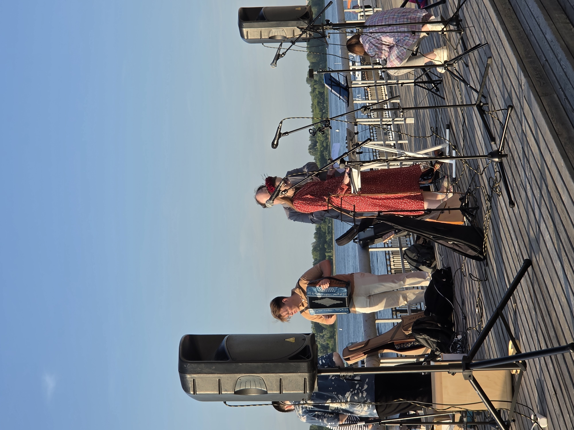 A band is performing on a wooden deck with a body of water in the background, possibly the Volga River in Nizhny Novgorod, Russia. The sky is clear and blue. There are two large speakers on stands, microphones, musical instruments including an accordion and a keyboard. A man is playing the accordion, a woman in a red dress is playing the keyboard and singing, and a person is crouched down in the background. There are also other people visible in the background. The overall atmosphere suggests a live outdoor music event.