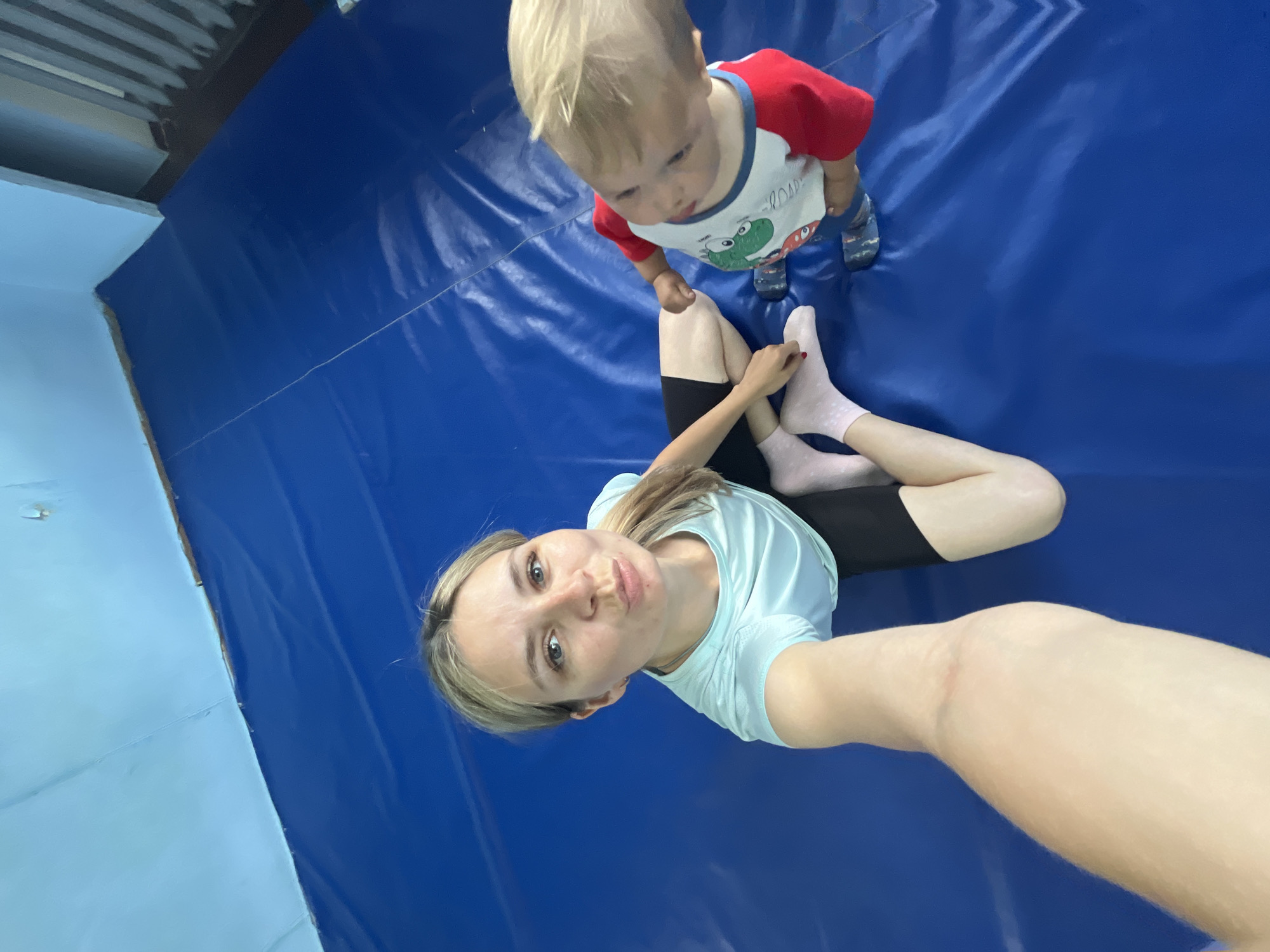 In this image, a woman and a young child are on a blue mat, likely in a gymnastics or play area. The woman is taking a selfie, holding the camera with her right hand. She has blonde hair and is wearing a light blue t-shirt and black shorts. Her left leg is bent, with her foot resting on the mat. She is looking at the camera with a playful expression, puckering her lips.
The child, who appears to be a toddler, is standing to the woman's right, looking down. He has blonde hair and is wearing a red and white t-shirt with a cartoon frog print and blue and grey patterned socks. The blue mat covers the floor and extends up the wall on the left side of the image, which is painted light blue. The ceiling above is made of corrugated metal, suggesting an indoor setting, possibly a sports facility. The lighting appears to be artificial. No specific time of day or weather conditions can be determined from the image. The scene depicts a casual, everyday moment between a mother and child.