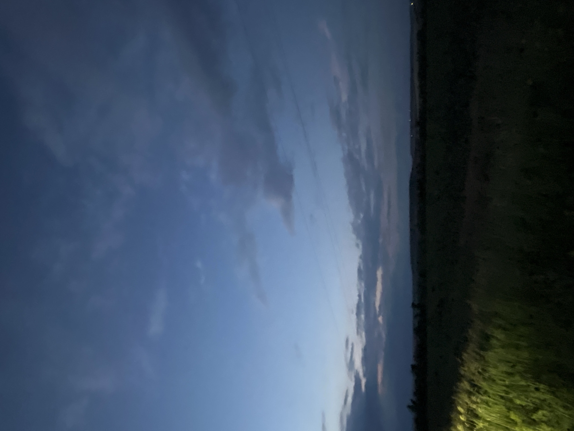 The image depicts a wide, open field at dusk in Poltavka, Ukraine. The sky is a deep blue with scattered, dark clouds, suggesting the end of daylight. A distant, dark horizon is visible, possibly indicating rolling hills or treelines. The foreground is dominated by a field of dark green grass or crops, with a bright, yellowish light illuminating the left side of the frame, suggesting a light source such as a vehicle headlight. The overall impression is of a rural landscape transitioning from day to night. There are no people or discernable actions taking place within the scene.