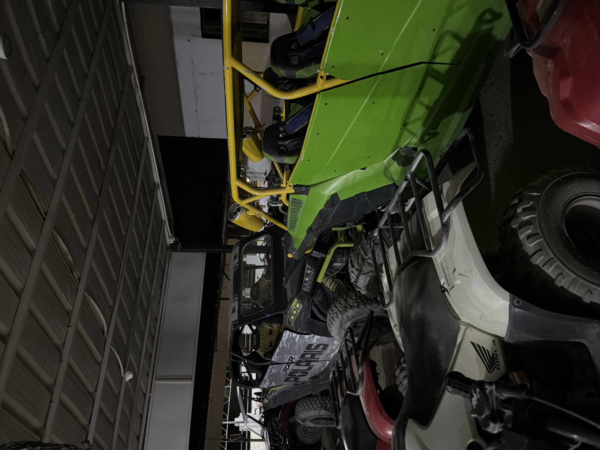 The image depicts an indoor storage area, likely a garage or workshop, in Sharm El Sheikh, Egypt. The scene is dimly lit, with the corrugated metal roof and supporting beams clearly visible overhead. Various all-terrain vehicles (ATVs) and utility task vehicles (UTVs) are parked closely together.
In the foreground, a white ATV, possibly a Honda given the visible logo, is parked on the left. A portion of a red ATV is visible on the right. Behind these, a black Polaris RZR UTV is prominent, with its "POLARIS" branding clearly visible. It has a roll cage and rugged tires. Adjacent to the Polaris, a bright green UTV with a yellow roll cage is parked, featuring multiple seats. Another dark-colored vehicle is partially visible behind the Polaris. The overall impression is one of recreational vehicles being stored. The setting appears functional rather than decorative, with exposed structural elements and dust or dirt visible on the vehicles and floor.