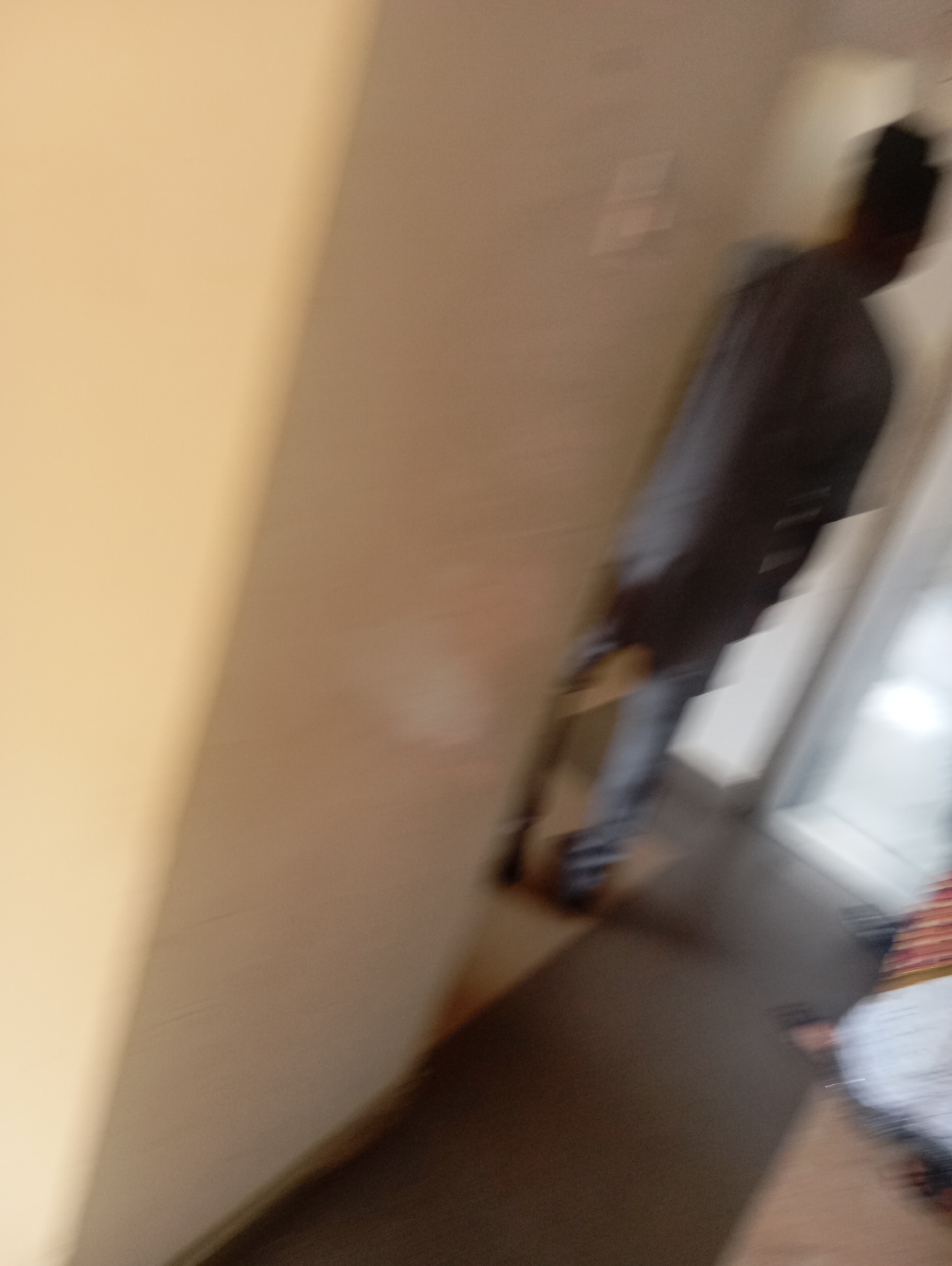 A motion-blurred image depicts a person standing in what appears to be an indoor setting. The person is dressed in dark clothing, possibly a long robe or coat, and is facing away from the camera. Their posture suggests they might be walking or leaning. The background is indistinct due to the blur, but it seems to be a hallway or corridor with neutral-colored walls and a dark floor. There is a white object, possibly a doorway or a partition, to the right of the person. The lighting is diffuse, suggesting it could be daytime, but without clear indicators like windows or shadows, the time of day is uncertain. The motion blur suggests movement during the capture of the image. No text is visible. The context of Jos, Nigeria, is provided but cannot be directly inferred from the visual content of the image itself.