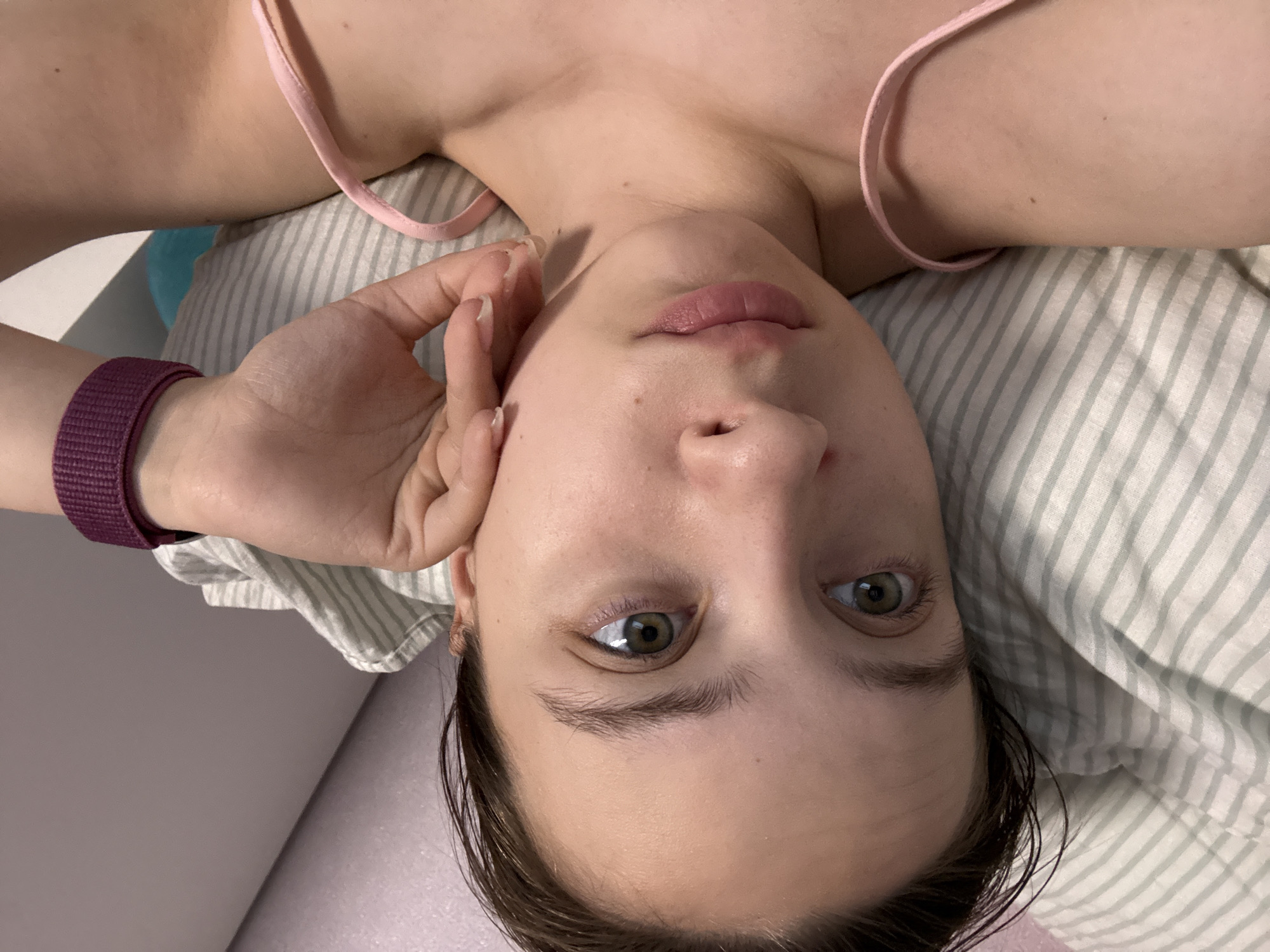 A woman lies on a striped pillow, wearing a pink tank top and a purple smartwatch. She touches her face with her left hand. The location is unknown; the reference to the city of Прикамский, Russia is irrelevant.