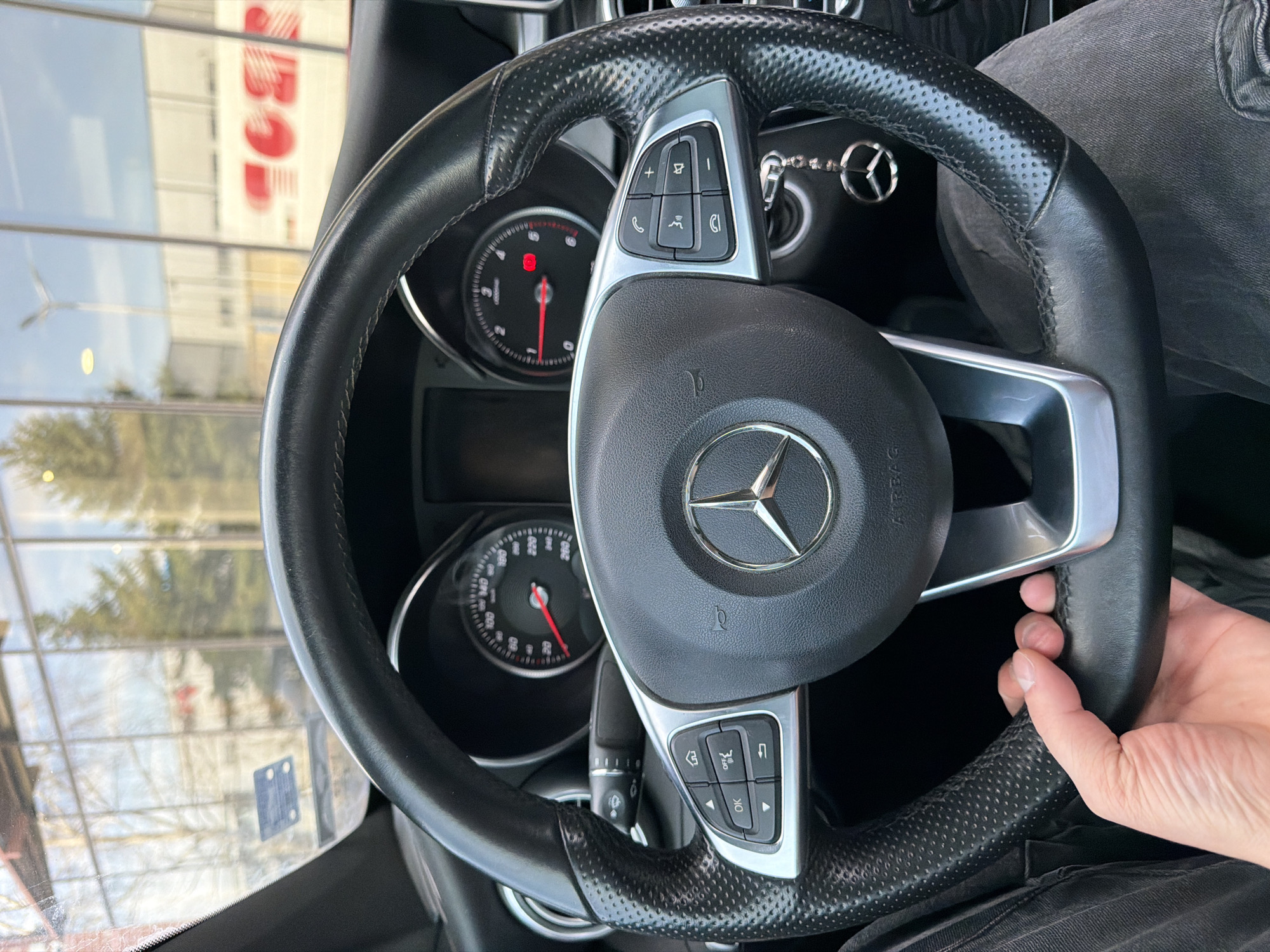 The image shows the interior of a Mercedes-Benz car, focusing on the steering wheel and dashboard. The steering wheel is black leather with perforated sections and silver-colored control buttons. The Mercedes-Benz logo is prominently displayed in the center. Behind the steering wheel, parts of the dashboard and instrument panel are visible, including the tachometer and speedometer. A hand is gripping the right side of the steering wheel. Through the windshield, a blurred view of the exterior can be seen. The red letters "TOR" are visible, part of a larger sign, suggesting the location is near or at a shopping center.
Given the signage and architectural style, the image was probably taken somewhere in Varna, Bulgaria.