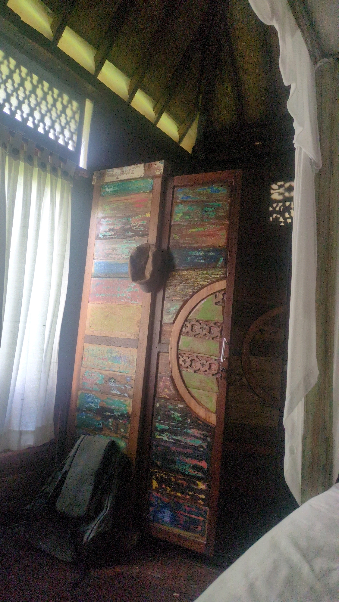 The image captures the interior of a room, likely a dwelling in Ubud District, Indonesia, with a distinct rustic and bohemian aesthetic. The focal point is a pair of tall, weathered wooden doors with a distressed, multi-colored finish, featuring horizontal planks in shades of green, blue, red, and yellow, suggesting recycled or salvaged materials. One door is slightly ajar, revealing a glimpse into a darker space.
Dominating the upper portion of the image is a thatched or wooden ceiling with visible rafters, contributing to the natural, tropical ambiance. Light streams in from a patterned window on the left, casting intricate shadows onto the wall and the door. A white, sheer curtain hangs across the window, diffusing the light and adding a soft texture.
In the lower left corner, a dark, canvas backpack rests on the floor, leaning against the door. A brown, fur-like object, possibly a hat or a decorative item, is affixed to the left door. To the right of the doors, a circular wooden element with an intricate carved design is partially visible, further enhancing the room's unique character. The floor appears to be a dark, polished wood or tile. A white fabric, possibly bedding or another curtain, is present in the lower right foreground, partially obscuring the view. The overall lighting is dim, with specific areas brightly illuminated by the sunlight.