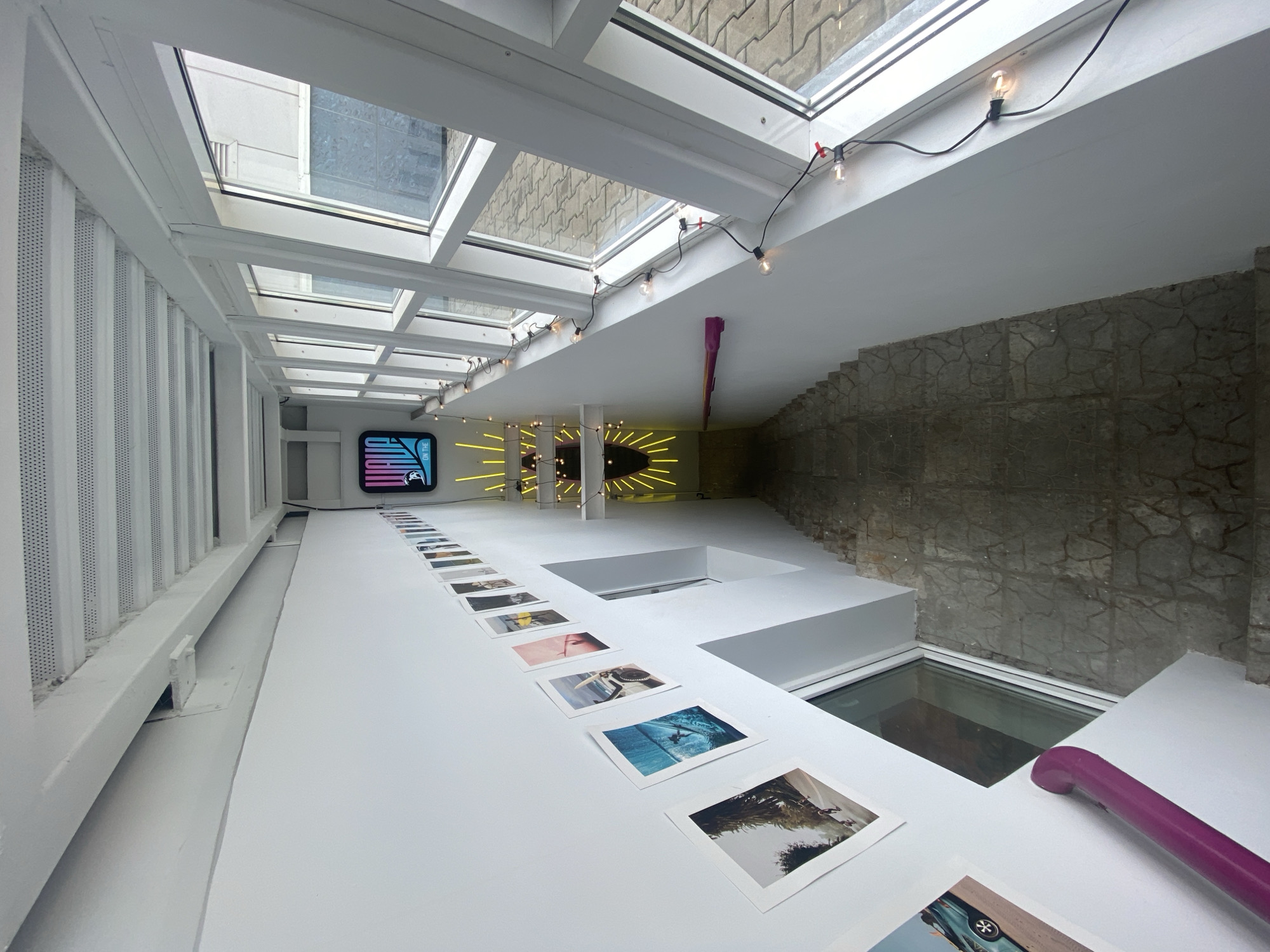 The image shows an indoor staircase with white walls and white ceiling beams. A row of framed photographs is mounted on the left wall, progressing down the stairs. A neon sign with the word "WAVE" in blue and pink, and a silhouette of a surfer, is visible at the top of the stairs. Yellow neon lights form a sunburst pattern on the wall below the sign. String lights with exposed bulbs are draped along the right wall, near a series of large windows that overlook an outdoor area with paving stones and some vegetation. A purple handrail runs along the left wall. The floor of the staircase is tiled with a gray, stone-like pattern. The scene appears to be a modern, interior space, possibly a cafe or art gallery, designed with a vibrant, surf-themed aesthetic. The location is identified as Khabarovsk, Russia, though no specific landmarks or contextual clues within the image directly confirm this location. There are no people visible in the image.