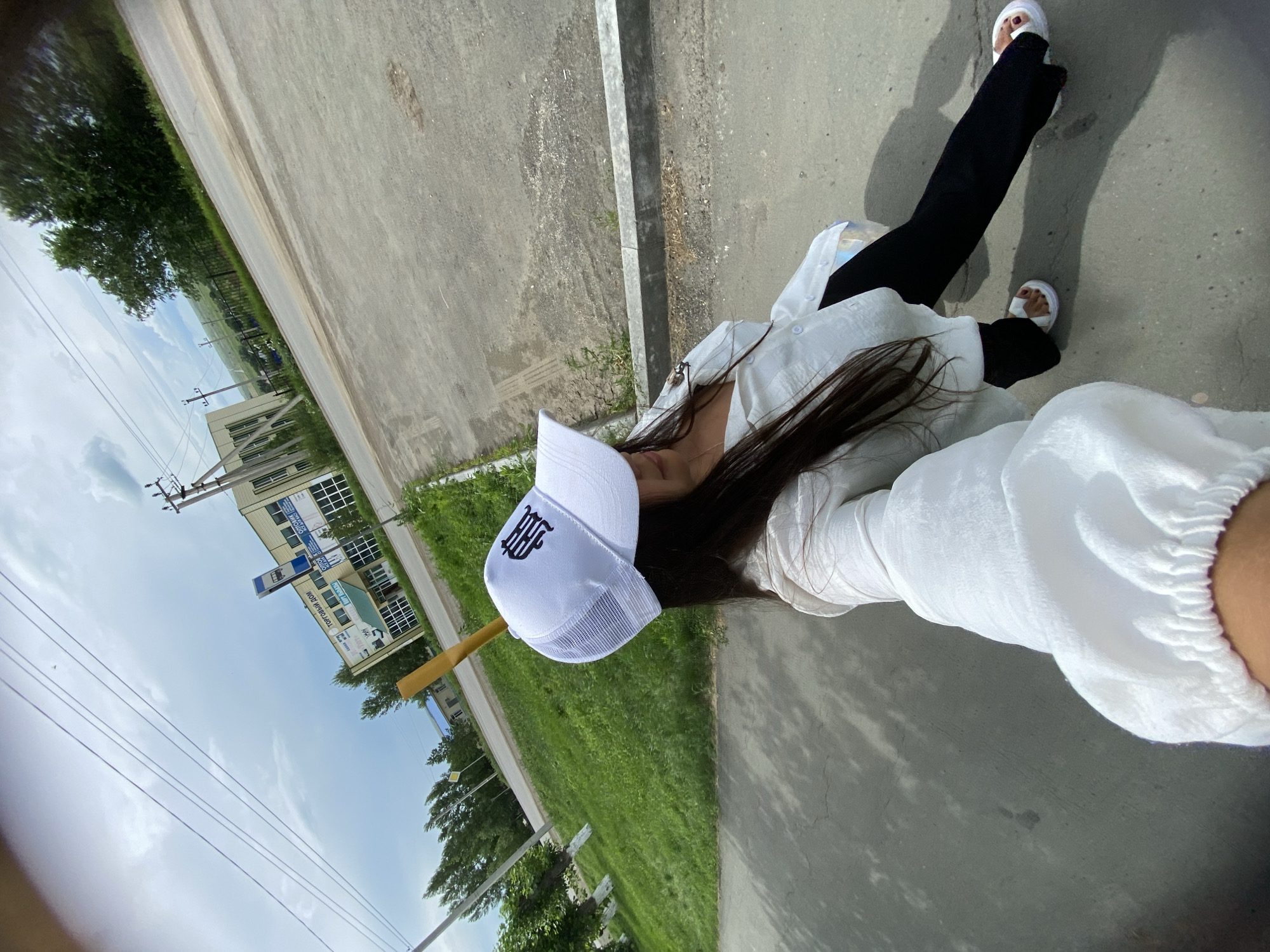 An outdoor, wide-angle, eye-level selfie is taken by a young woman wearing a white baseball cap with a black emblem, a white long-sleeved shirt, and black pants. She is standing on a paved area, possibly a sidewalk or road, with a curb to her right. In the background, a two-story building with a sign that reads "ТОРГОВЫЙ ДОМ" (Trading House) is visible, along with utility poles and trees. Beyond the building, a green, hilly landscape stretches under a cloudy sky. The scene appears to be in the city of Akyar, Russia, given the signage. The woman is holding the camera with her right hand, her arm extended.