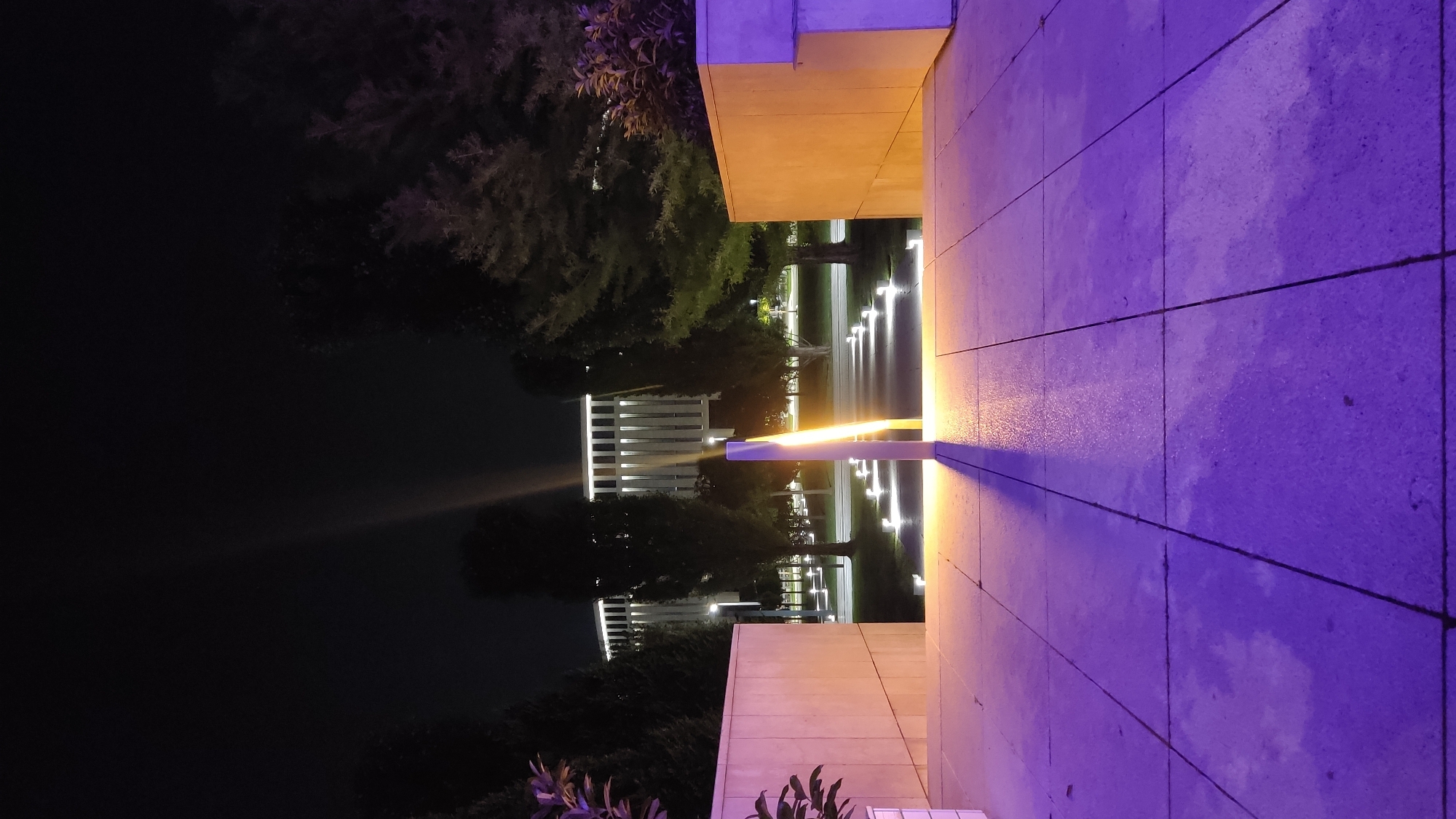The image depicts an outdoor scene at night, likely a park or public space, illuminated by artificial lights. In the foreground, a paved walkway with visible grout lines is bathed in purple light, with some areas reflecting the light more intensely, suggesting a wet surface. A tall, slender pillar stands near the center of the walkway, emitting a bright yellow light that casts a long shadow onto the pavement.
Beyond the foreground, a pathway lined with small, rectangular lights recedes into the distance. Flanking this pathway are several trees with dark foliage and a long, low, cream-colored wall on the left, and a similar wall on the right which is shorter and has a lighter colored vertical section at its far end.
In the background, a large, modern building with a facade of vertical, white elements and illuminated horizontal bands is visible between the trees. The sky is dark, indicating nighttime. The overall impression is one of a well-lit, possibly tranquil, urban nightscape. There are no people or animals visible in the image.