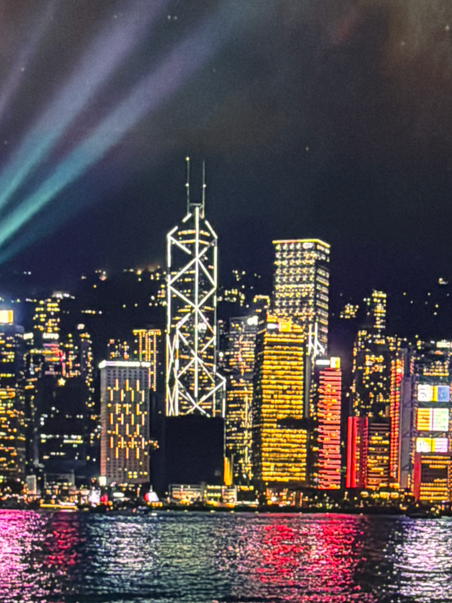 The image displays a nighttime cityscape, likely of Hong Kong, characterized by numerous illuminated skyscrapers and a body of water in the foreground reflecting the city lights. Prominent beams of light, possibly from a laser show or searchlights, cut through the dark sky from the upper left. The most distinctive building in the center is a tall skyscraper with a white, angular lattice structure on its exterior. Surrounding it are other buildings of varying heights, all aglow with yellow and orange lights. The water exhibits a vibrant reflection of pink and red lights from the city. There are no people visible in the image. The provided image does not depict Kafr Yasif, Israel.