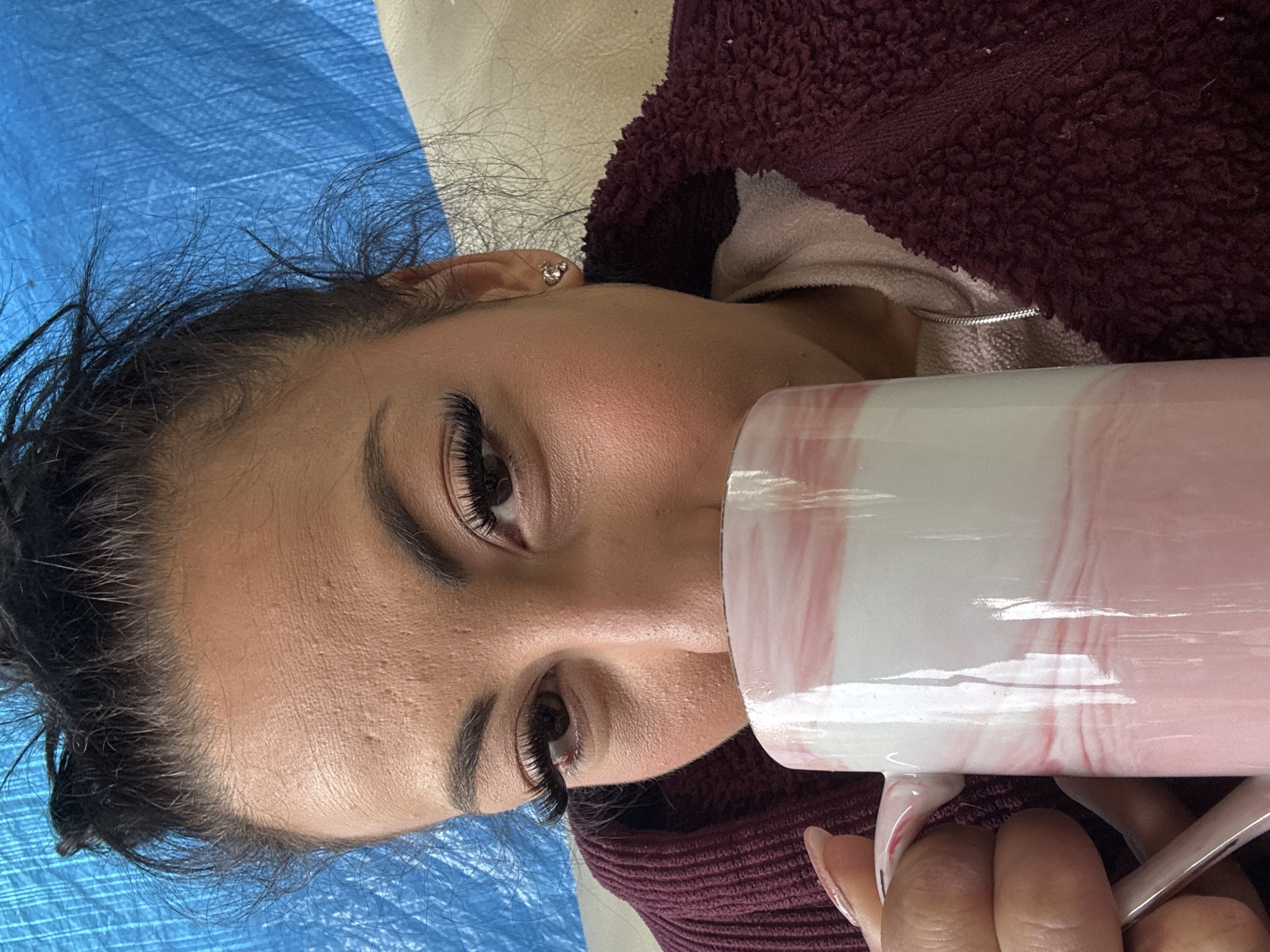 A woman lies on a light-colored surface covered with a blue sheet or cloth. She is wearing a burgundy-colored, textured, hooded garment over a light-colored top. She has dark hair and noticeable eyelash extensions. She holds a large white mug with pink marble swirl designs in front of her face with her right hand, where the manicured fingers with nail extensions are visible. The photograph does not contain location information about Warrington, United Kingdom.