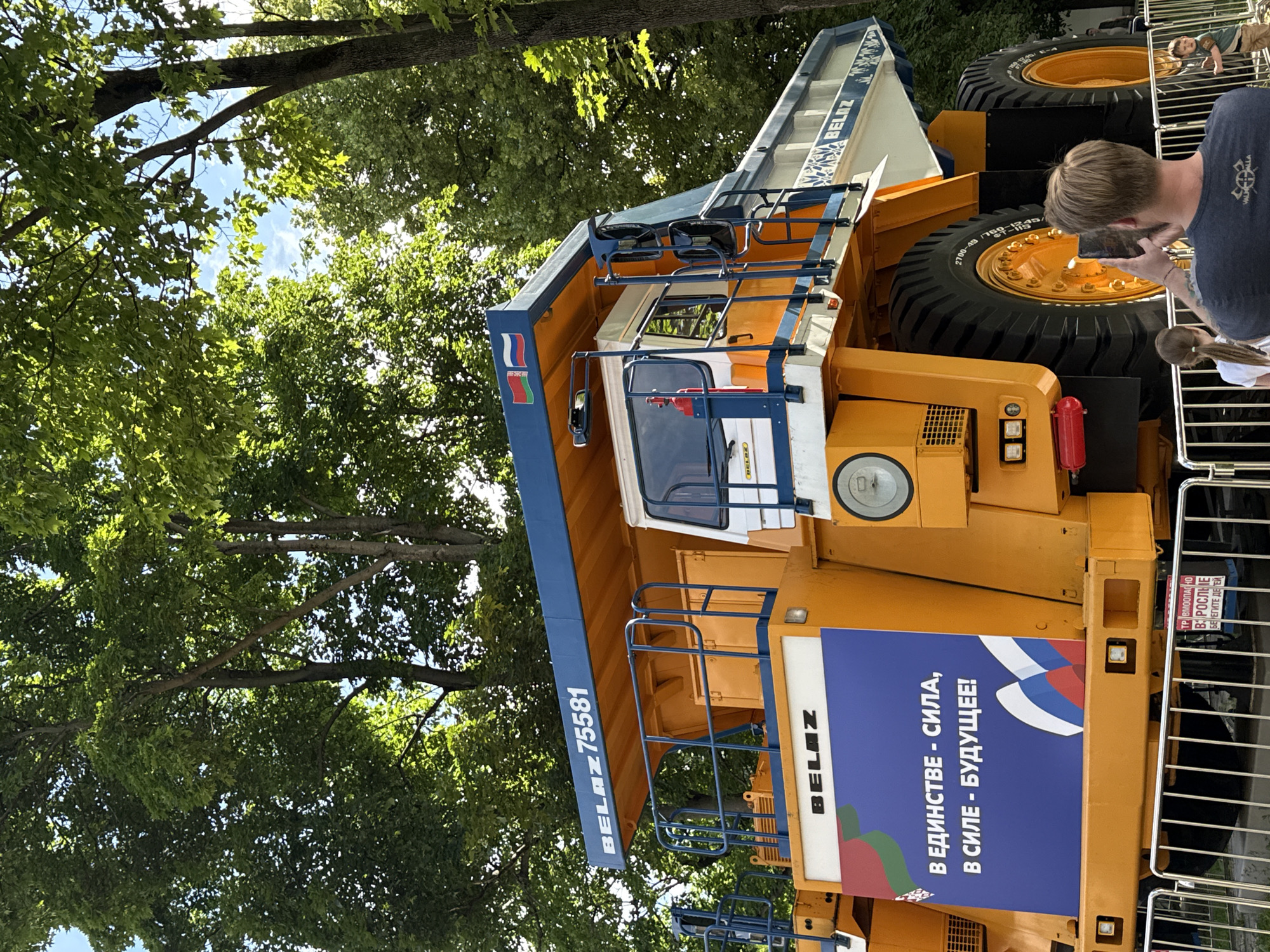 This media file shows a large yellow and blue Belaz mining truck parked outdoors, likely on a sunny day given the bright lighting and dappled shade from overhead trees. The truck is the central object, with its massive tires and rugged construction dominating the frame.
Several people are visible around the truck, interacting with it and each other. A man in a dark t-shirt is in the foreground, holding a phone up as if taking a picture or video of the truck. A young child in a light-colored shirt and shorts is seen lying on the truck's tire, looking upwards. Another child with dark hair is partially visible behind a metal railing. The presence of people suggests this might be an exhibition, a public event, or a special viewing of the truck.
The truck itself has various labels. The words "BELAZ 75581" are prominently displayed on its side. Another label reads "BELAZ" vertically. A large blue banner on the side of the truck features Russian text that translates to "In unity - strength, in strength - the future!" and includes a depiction of the Russian flag. A smaller red sign with white text appears to warn of danger, stating "TRAUMA HAZARDOUS. PROTECT CHILDREN."
The setting appears to be a park or open area with mature trees providing shade, suggesting it is a pleasant, likely daytime event. The overall mood seems to be one of curiosity and admiration for the impressive machinery.