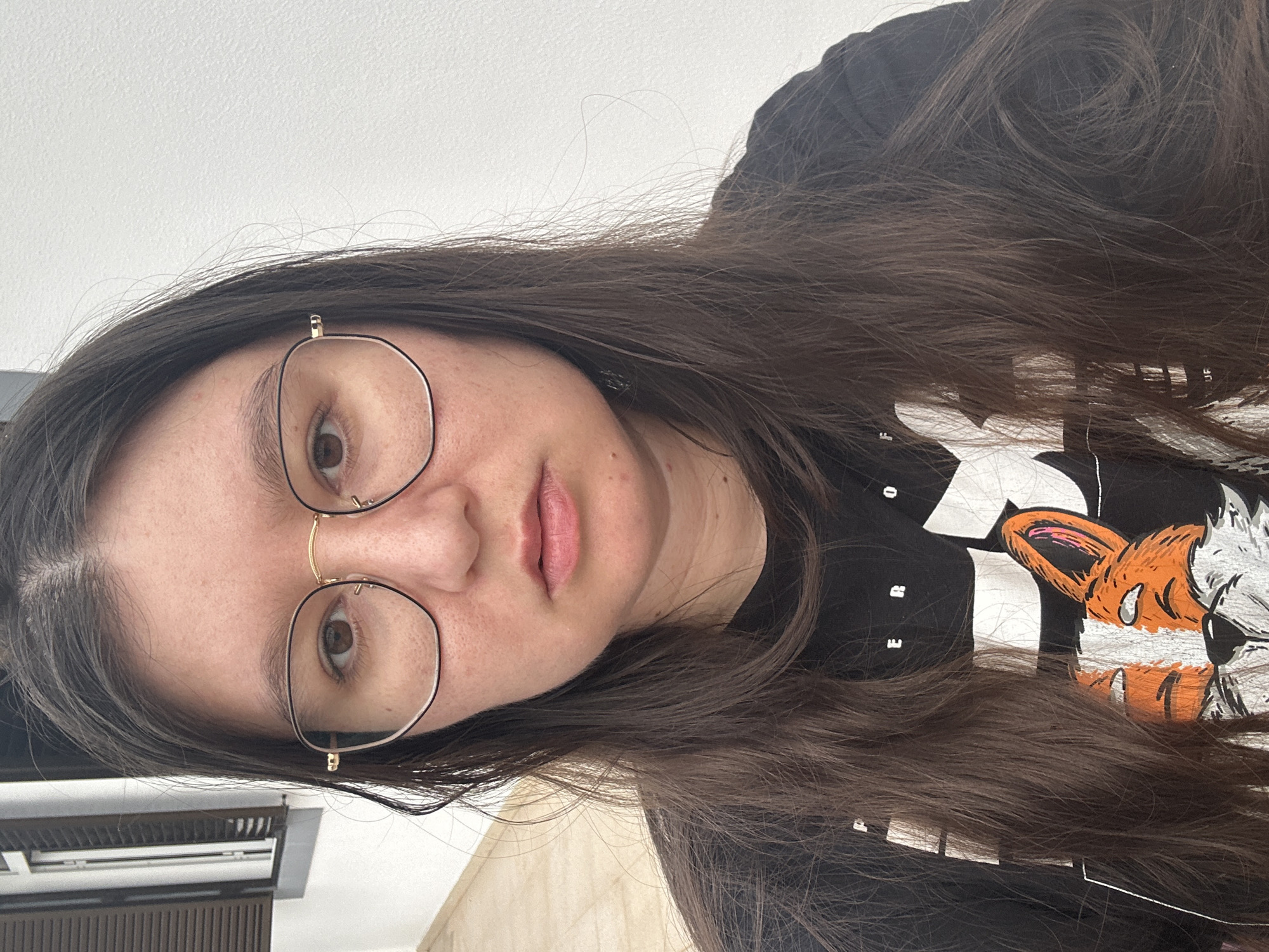 A woman with long brown hair and glasses is facing the camera. She is wearing a black T-shirt with a graphic design featuring a fox and lettering. The background includes a partially visible window with slats and a light-colored wall. The location appears to be indoors, possibly in Câmara de Lobos, Portugal.