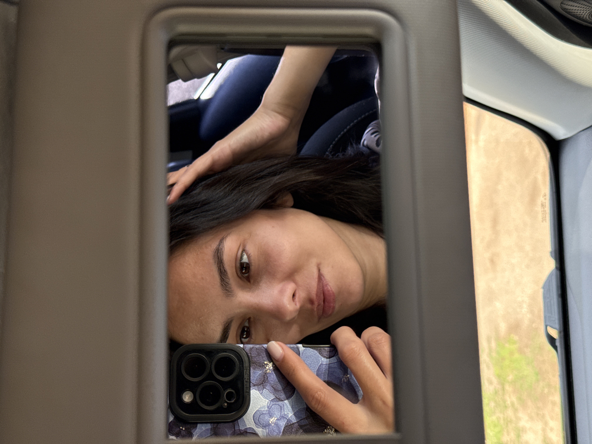 A woman is taking a selfie in a car's visor mirror. She is holding a smartphone with a floral-patterned case in her left hand, and her right hand is touching her hair. The mirror reflects her face, her left hand with the phone, and her right arm and hand. In the background of the reflection, a portion of a car's interior, including a dark-colored seat, is visible. The car appears to be parked or moving slowly, as indicated by the blurred outdoor scenery of dry grass and a dirt road reflected in the lower part of the visor. The image is taken from a slightly downward angle, looking up at the visor. The exact location in Chaikovsky, Russia, cannot be determined from the visual information provided.