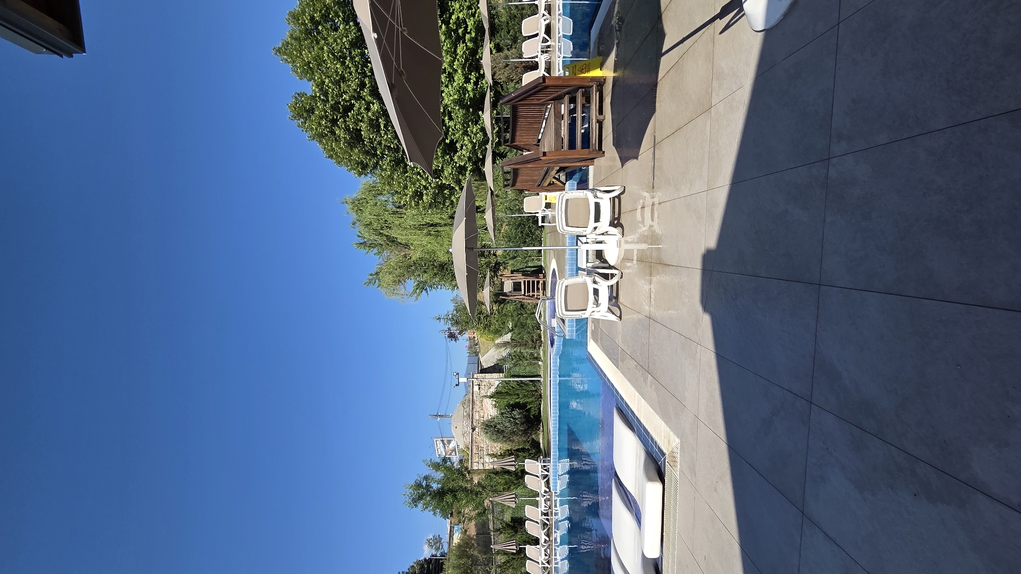 The image shows an outdoor swimming pool area on a sunny day. The pool is filled with clear blue water and is surrounded by a tiled patio. Several lounge chairs and umbrellas are placed around the pool, some unoccupied. A wooden deck with stairs leads into a section of the pool. In the background, there are lush green trees and a clear blue sky. Beyond the trees, there appears to be a small cluster of buildings with domed roofs, suggesting a historical or cultural element to the location. The scene evokes a sense of relaxation and leisure, typical of a resort or hotel. The specific location in the city of Bania, Bulgaria, is not explicitly indicated by any visual cues within the image itself, but it is implied by the prompt.