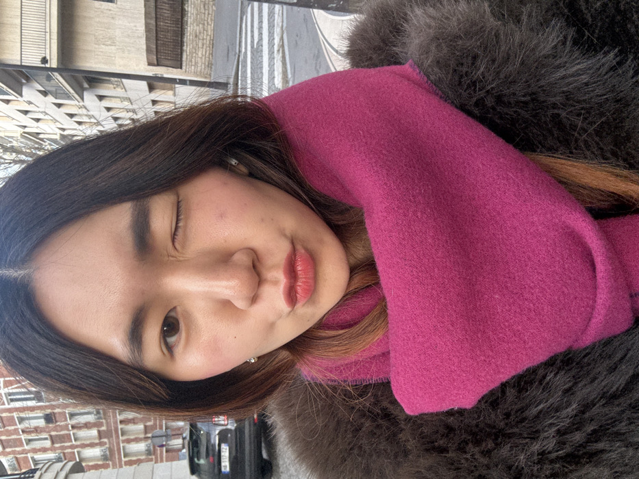 A young East Asian female is depicted in a close-up, selfie-style photograph taken on a street in Paris, France. She has long, dark brown hair with lighter reddish-brown undertones, centrally parted. She is wearing a dark brown, possibly fur-trimmed, coat and a vibrant magenta scarf wrapped around her neck and chest. Her left eye is closed in a wink, while her right eye is open, revealing a brown iris. Her lips are pursed in a slight pout, accented by coral-red lipstick. Small stud earrings are visible in her ears, and a rosy blush is applied to her cheeks.
The background indicates an urban street scene. To the subject's left, a multi-story building with a reddish-brown brick facade is partially visible. A dark-colored SUV, bearing a French license plate "HA-312-YZ", is parked on a cobblestone street. To the subject's right, a lighter-colored multi-story building with multiple balconies can be seen. A paved street with a white pedestrian zebra crossing is also present, suggesting a public roadway. The lighting indicates daylight, with no harsh shadows.
