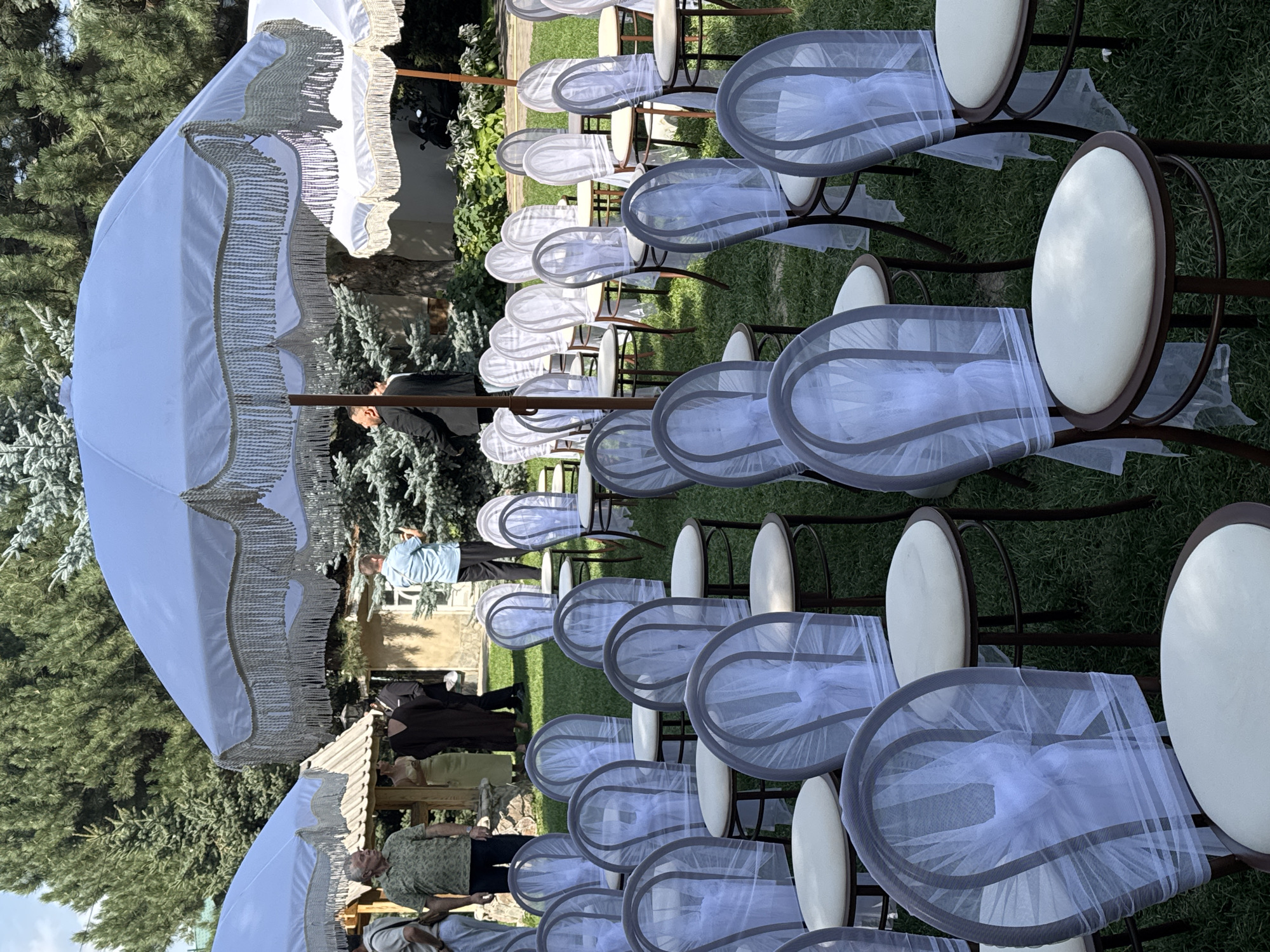 The image depicts an outdoor wedding ceremony setup in Koy Tash, Kyrgyzstan. In the foreground, multiple rows of chairs, each adorned with white sheer fabric tied around the back, are arranged on a grassy lawn. These chairs are light brown with tan padded seats. In the middle ground, several large, white, fringed umbrellas with wooden poles provide shade over the seating area. Behind the umbrellas, a few people are visible. To the left, three men stand, one in a green collared shirt and jeans, another in a light blue shirt and dark pants, and a third in a dark suit. Further back, a woman in a light yellow dress and another in a dark brown dress are partially visible. The background consists of trees, including coniferous trees, and a building with a light-colored facade and a wooden roof. The lighting suggests it is daytime, with sunlight casting shadows.