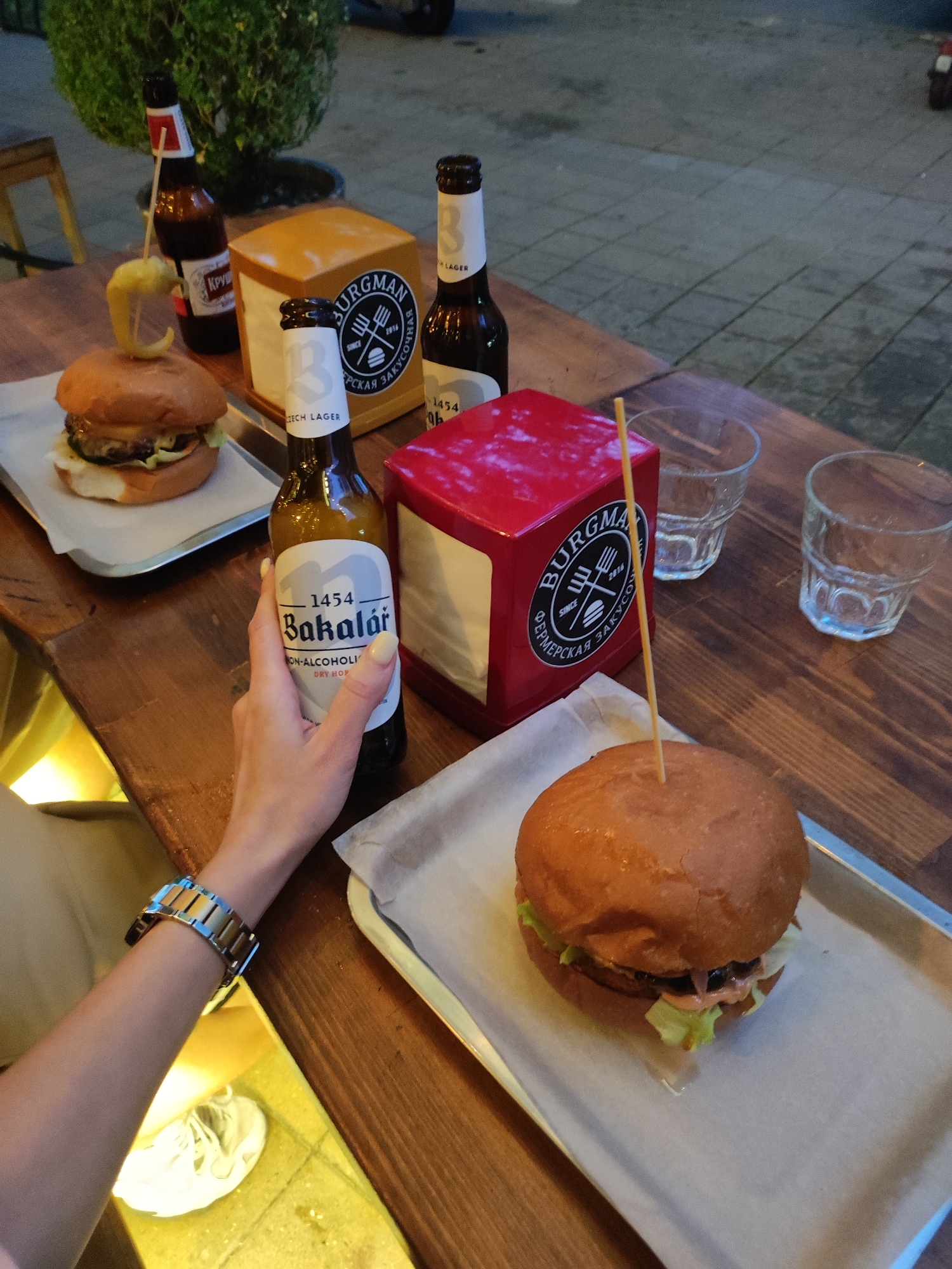 The image shows a wooden table in an outdoor setting, possibly a restaurant, with food and drinks. Two burgers are placed on metal trays, each secured with a toothpick. Two beer bottles and two empty glasses are also on the table. Napkins in a red and yellow holder with the "Burgman" logo are positioned between the items. A hand is holding a bottle of "Bakalář" non-alcoholic beer. The scene is set in the city of Sochi, Russia.