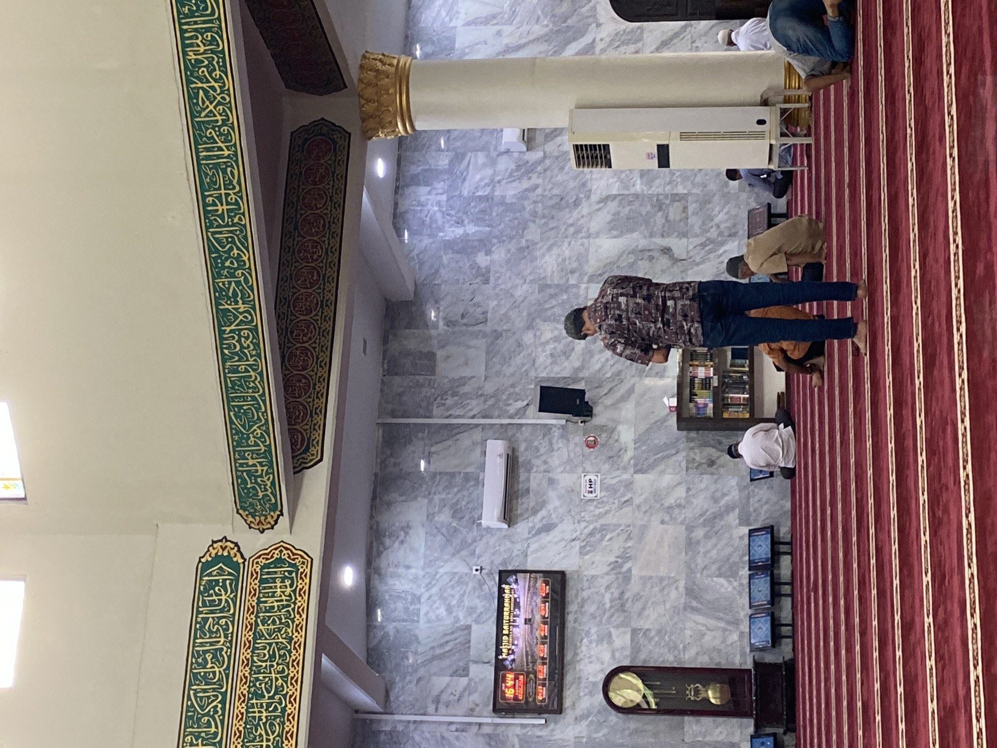 The image depicts the interior of a mosque, specifically Masjid Baiturrahman in Lhokseumawe, Indonesia. The foreground features a red carpet with white stripes, indicating rows for prayer. Several individuals are engaged in prayer, either standing or kneeling. In the midground, a man in a patterned shirt and jeans stands facing away from the camera, appearing to be praying. To his left, a digital clock and prayer time display is mounted on a marble-patterned wall. Further to the left, a grandfather clock is visible. The walls are adorned with green and gold Islamic calligraphy. The background shows more individuals praying, a bookshelf, and air conditioning units. A prominent white pillar with a golden capital stands to the right of the frame. The overall environment is that of a religious gathering place during prayer.