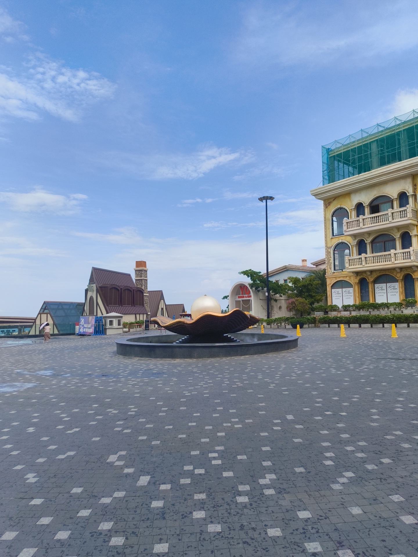 The image depicts a public square in Phu Quoc, Vietnam, featuring a large fountain with a golden shell-like structure topped with a sphere. The square is paved with dark grey square tiles interspersed with lighter square tiles. In the background, there are several buildings with distinct architectural styles. To the left, a building with a brown, sloped roof and white trim resembles a European-style church or lodge. Adjacent to it is a modern structure with a glass facade. To the right, a cream-colored, multi-story building with arched windows and balconies is under construction, covered with green netting. A lamppost stands in the center of the square, between the fountain and the buildings. A single person is visible walking on the left side of the square, holding a white umbrella. Several yellow traffic cones are placed along the edge of the fountain area. The sky is blue with scattered white clouds.