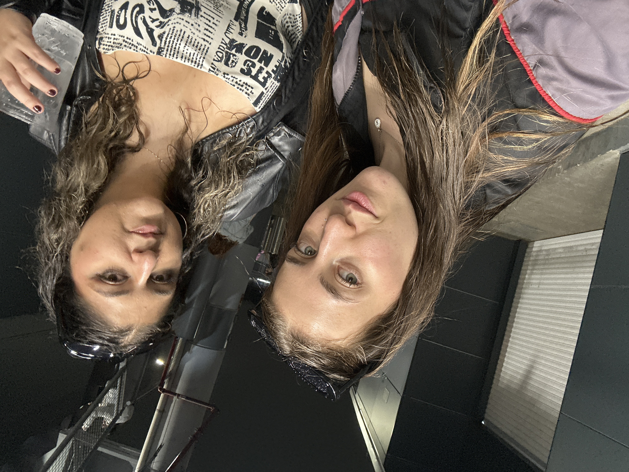 Two people are pictured indoors. The person on the left has dark hair with curly, light brown highlights, and is wearing a black leather jacket over a white top with black print, and is holding a plastic cup with a clear liquid inside. The person on the right has long brown hair, is wearing a black jacket, a silver necklace, and has a stud piercing on their nose. The background shows interior architectural elements including a corrugated metal panel and dark colored panels. It is not possible to determine the location of the photo within Mexico City, Mexico based solely on this image.