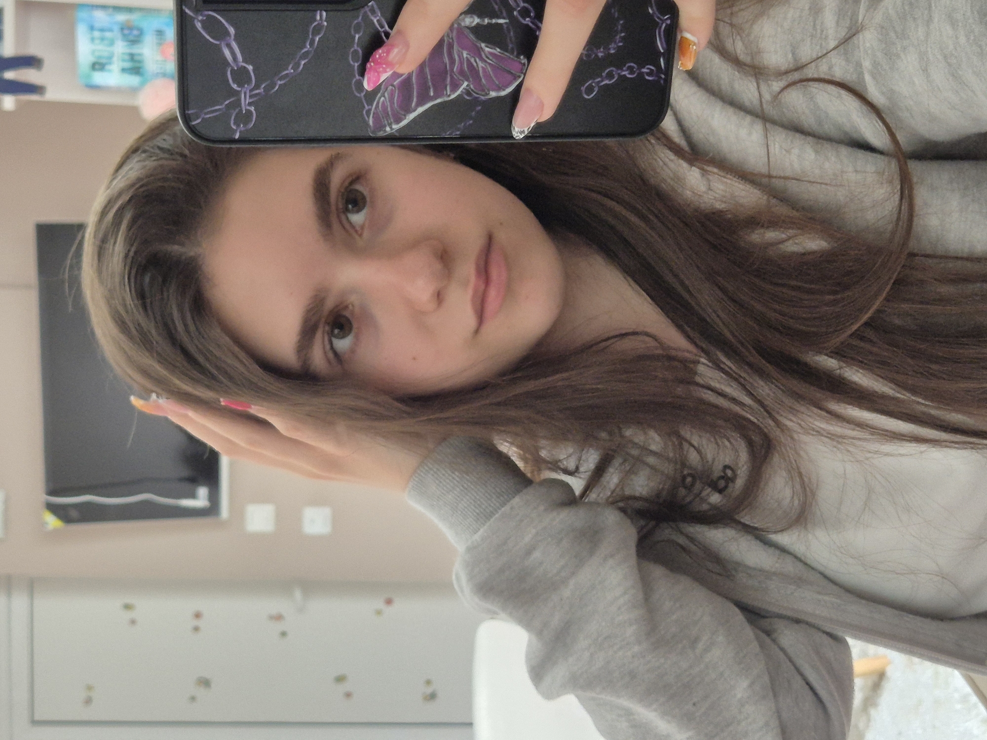 A young woman poses for a selfie, holding a black smartphone with a decorated case. Her hair is brown and falls around her face and shoulders. She is wearing a grey sweatshirt. The background includes a wall with a television mounted on it, and a whiteboard-like surface with small colored spots. The location is Pavlikeni, Bulgaria.