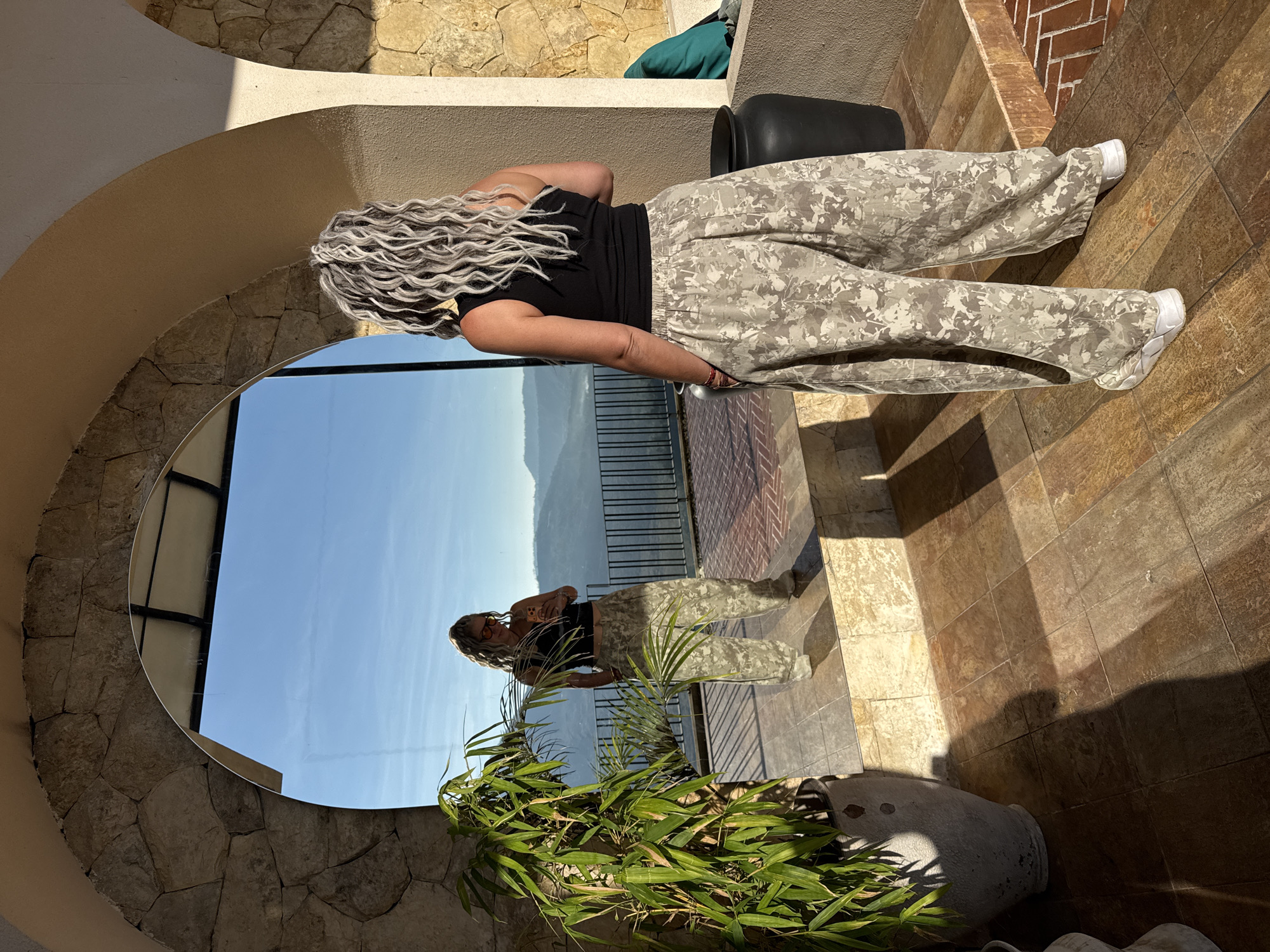 The image depicts an outdoor or semi-outdoor space in Kintamani, Indonesia, featuring a woman standing with her back to the camera, interacting with her reflection in a large arched mirror. The setting is characterized by stone and stucco architectural elements and an expansive mountainous landscape.
The central figure is an adult female with long, curly, predominantly grey and silver hair, wearing a black sleeveless crop top and loose-fitting, wide-leg pants with a light, abstract camouflage or leaf pattern in muted beige and grey tones. She wears white sneakers. Her reflection in the mirror, positioned to the left, shows her holding an orange-cased smartphone in her right hand, suggesting she is taking a mirror selfie or checking her device. In her reflection, she is also wearing orange-tinted sunglasses.
The mirror is set within a robust archway constructed of irregular, rough-hewn stone blocks. To the left of the mirror, a tall, leafy green plant, possibly bamboo, grows out of a large, textured grey pot. The floor is covered in square tiles of varying earthy tones, including brown, beige, and reddish hues. Strong, directional sunlight casts long shadows across the tiled floor, indicating either early morning or late afternoon.
Beyond the mirror and to the right of the woman, a vast natural landscape unfolds under a clear blue sky. This view includes multiple layers of rolling hills and mountains, consistent with the volcanic terrain of Kintamani. A dark metal railing is visible along the edge of the architectural structure, overlooking this vista. To the immediate right of the woman, a large, dark, cylindrical planter or pot stands on the tiled floor. The space is defined by an arched passage on the right with stuccoed walls and a stone base.