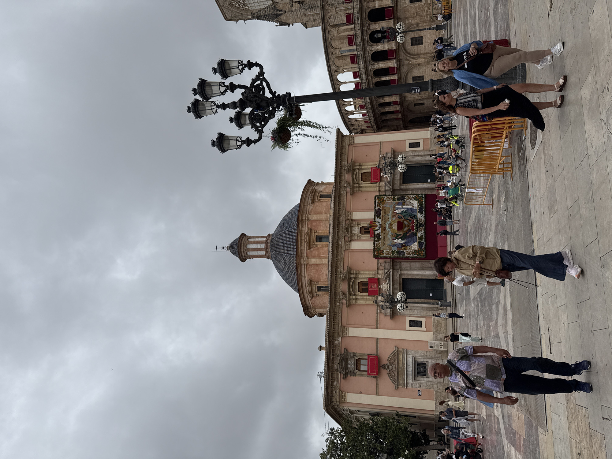 The image captures a public square in València, Spain, under an overcast sky, suggesting daytime without bright sunlight. The scene is rich with historic European architecture and shows various people engaged in casual activities.
At the center, a prominent pinkish-orange building with a large dome and a small cupola on top stands out. This building is the Basilica de la Mare de Déu dels Desamparats. Its facade features several red rectangular banners draped from windows and a large, colorful tapestry depicting a religious scene. Visible text on this tapestry includes "2023 ANIVERSARI" and "CORPUS CHRISTI". To the upper right of the basilica, partially obscured by an ornate black street lamp adorned with a hanging basket of greenery, is another building with arched windows and red shutters. A fragment of a Gothic-style stone building is visible in the upper left background.
The plaza floor is paved with light-colored stones. Several people are scattered across the square. In the mid-ground on the right, two women are standing near yellow crowd control barriers. One wears a black top, a dark skirt, and sandals, while the other is dressed in a black t-shirt, a blue jacket, and light pants with sneakers. Closer to the foreground, a man in a patterned short-sleeved shirt and dark trousers walks towards the viewer. Further back, on the left side of the square, another woman in a tan jacket and dark pants is visible. In the background, other individuals can be seen, some seated on chairs behind additional yellow barriers, suggesting the presence or preparation for an event or gathering. The overall impression is that of a typical day in a historic, active city square.