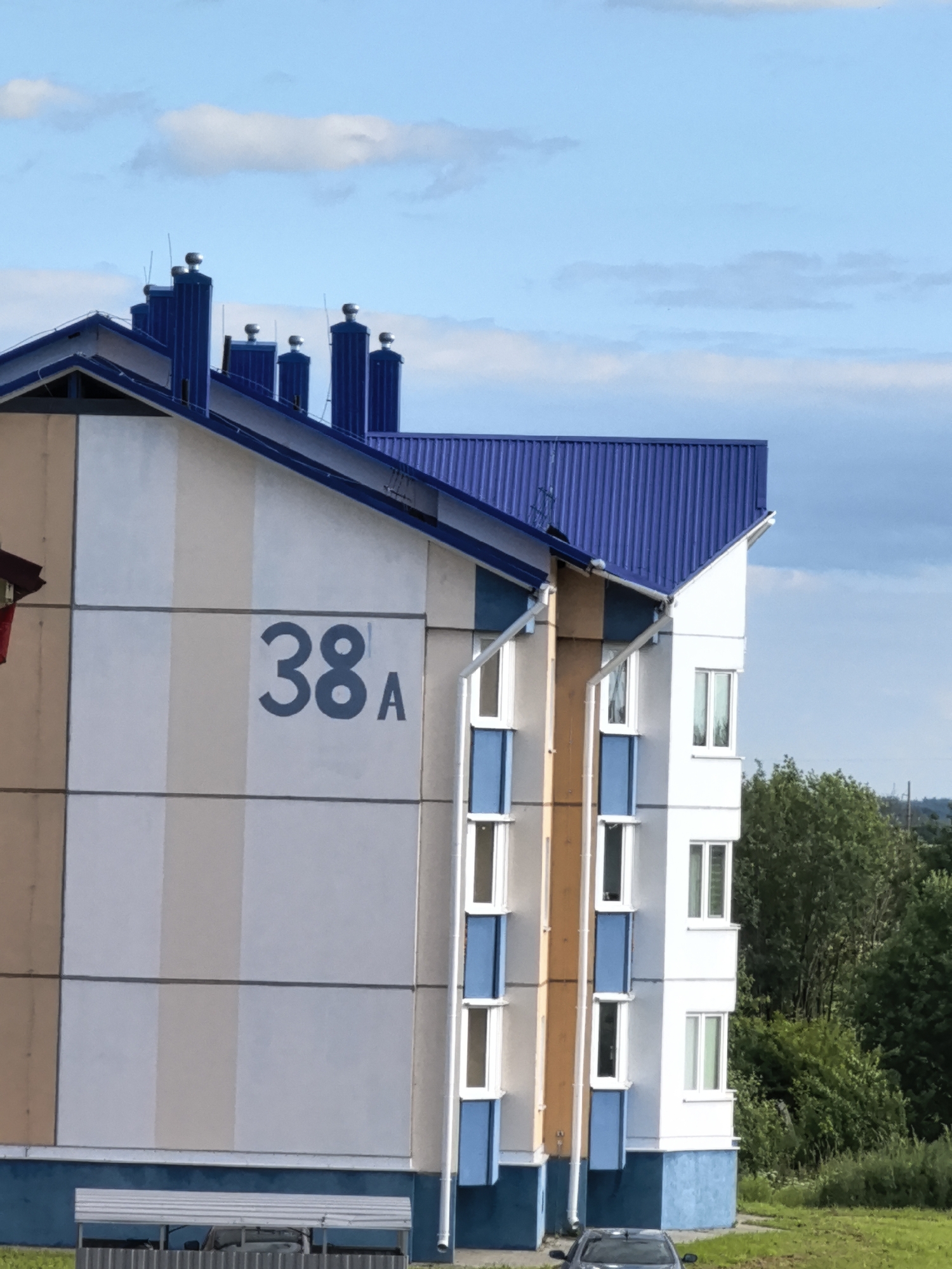 A side view of a multi-story building with a blue metal roof is shown against a partially cloudy sky. The building has a light beige and white facade with vertical blue and brown stripes. The number "38A" is prominently displayed in dark blue on the facade. Several blue chimney pipes with silver caps are visible on the roof. The building has multiple white-framed windows, some with blue trim. In the foreground, a white bench with a corrugated metal roof structure is visible, and a dark-colored car is parked on a grassy area to the right. Lush green trees and foliage are visible in the background.