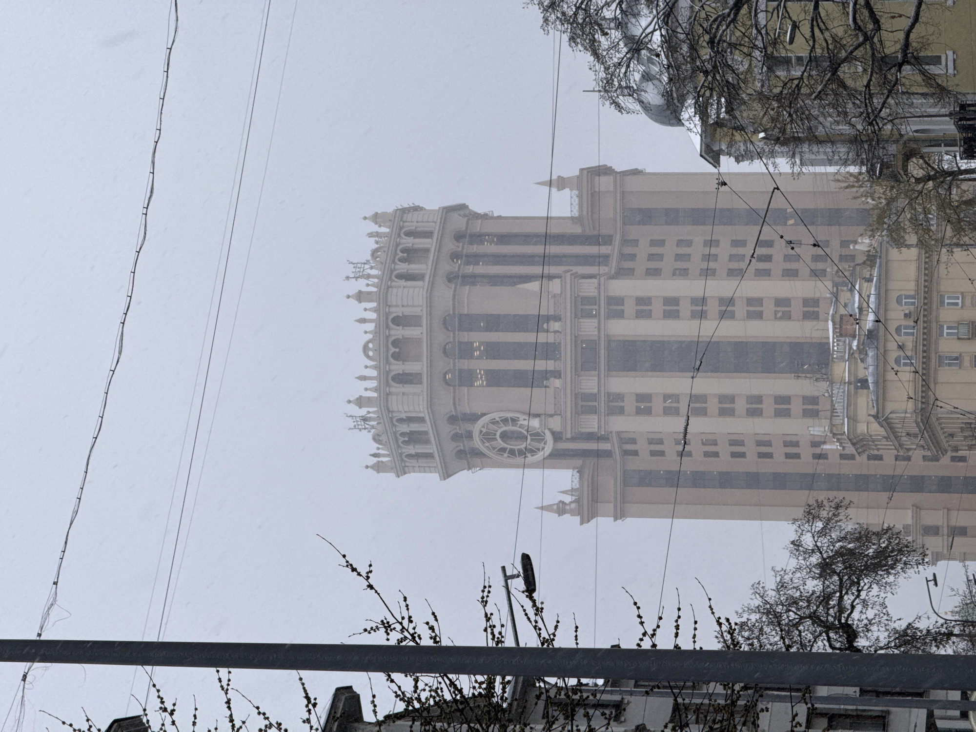 The image shows a building in Moscow, Russia, during snowy weather. The building is a high-rise with a distinctive architectural style, featuring a clock on its facade. Overhead electrical wires are visible in the foreground, along with bare branches. In the background, other buildings and trees are present. The sky is overcast and the environment appears to be urban.