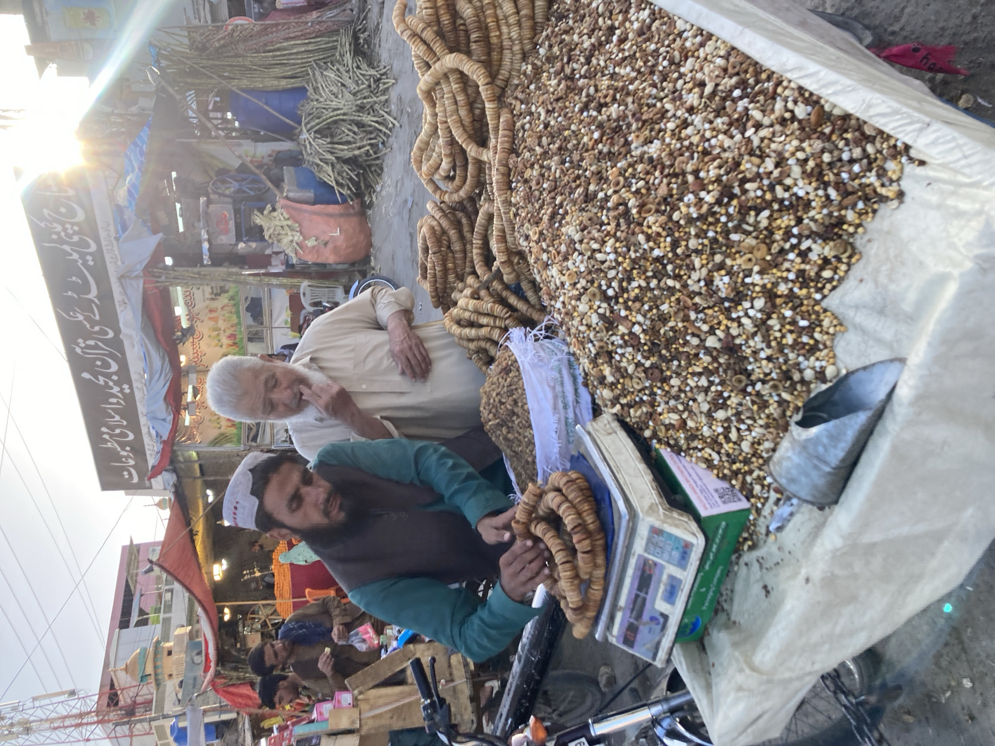 The image depicts a street scene, likely a market, in Quetta City Tehsil, Pakistan. A vendor, wearing a white cap and a green shirt over a vest, is weighing what appears to be dried figs using a digital scale. An older man with white hair stands next to him. A large bag containing a mixture of dried fruits and nuts sits nearby. Strings of dried figs are also visible. In the background, there are other vendors and shoppers, as well as sugarcane stalks. Street signs and structures consistent with an urban environment are seen.