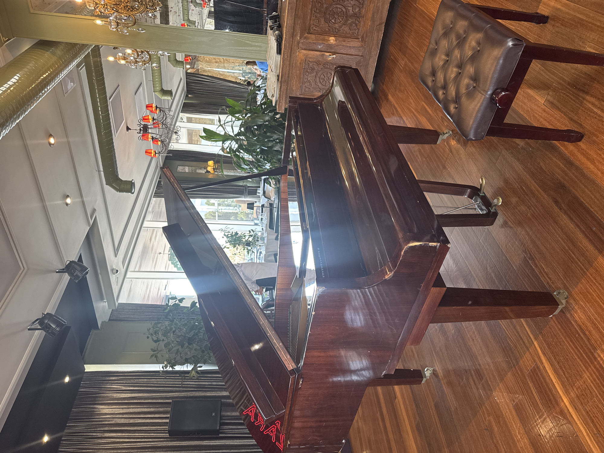 In the Sabail Raion, Azerbaijan, a dimly lit, upscale establishment features a polished grand piano as the central element. The piano, made of dark, reflective wood, has its lid partially open, revealing its interior. A tufted, dark brown piano bench sits to the right of the instrument. The floor is covered in rich, dark wood planks. In the background, draped curtains, a large speaker, and a wooden cabinet with intricate carvings can be seen. The ambient lighting includes decorative chandeliers with red accents and spotlights mounted on the ceiling. The environment appears to be a dining or event space, with tables and chairs visible through large windows in the distance, suggesting a restaurant or ballroom setting.