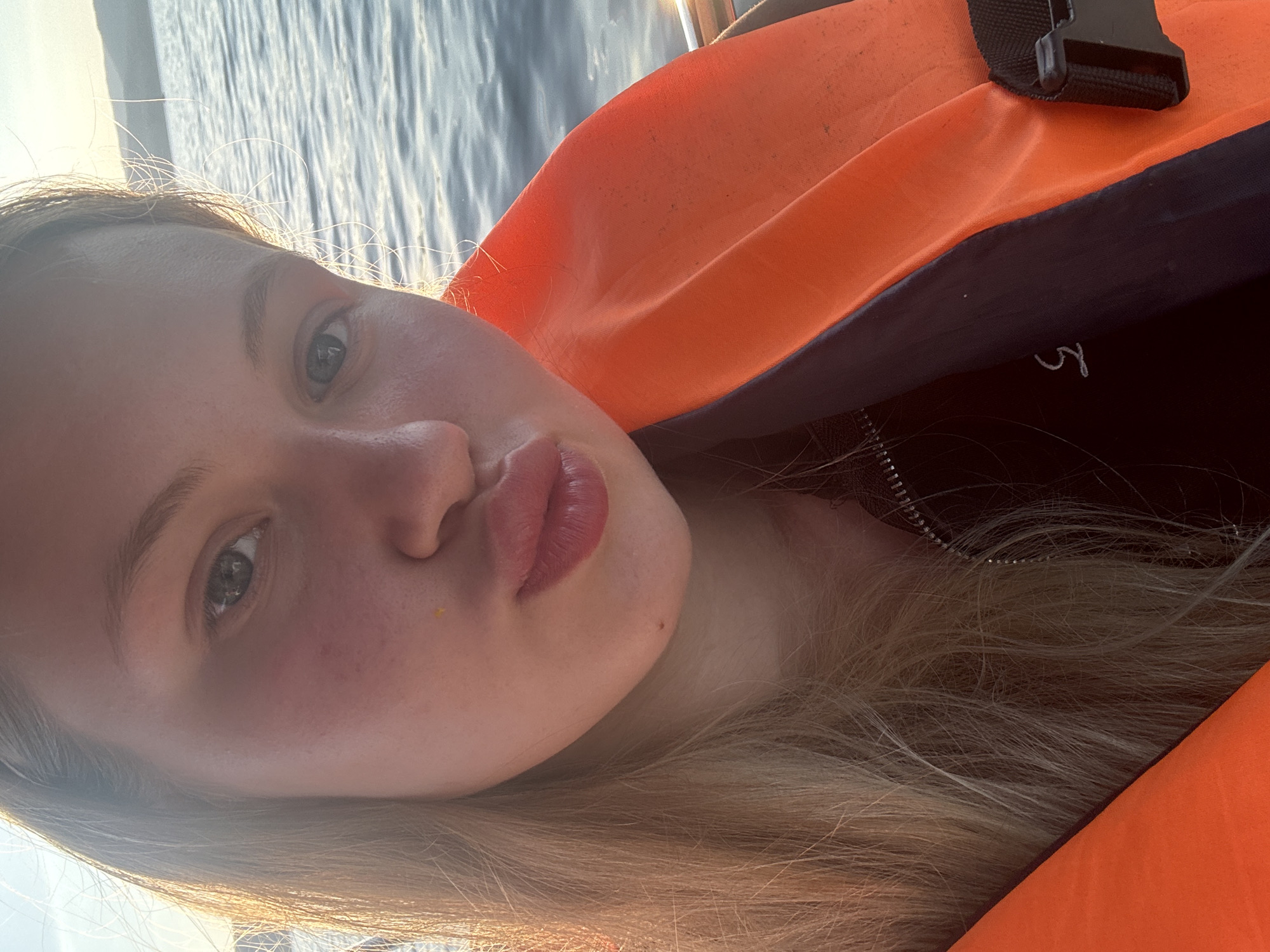 A young woman with blonde hair and a life jacket is taking a selfie on a body of water. The sun is setting, casting a warm glow on her face. She is pouting her lips as if to blow a kiss. The water is calm and reflects the light of the setting sun. In the background, hills can be seen on the horizon. The woman appears to be in good spirits and enjoying her time on the water. The location is the city of Turgoyak, Russia.