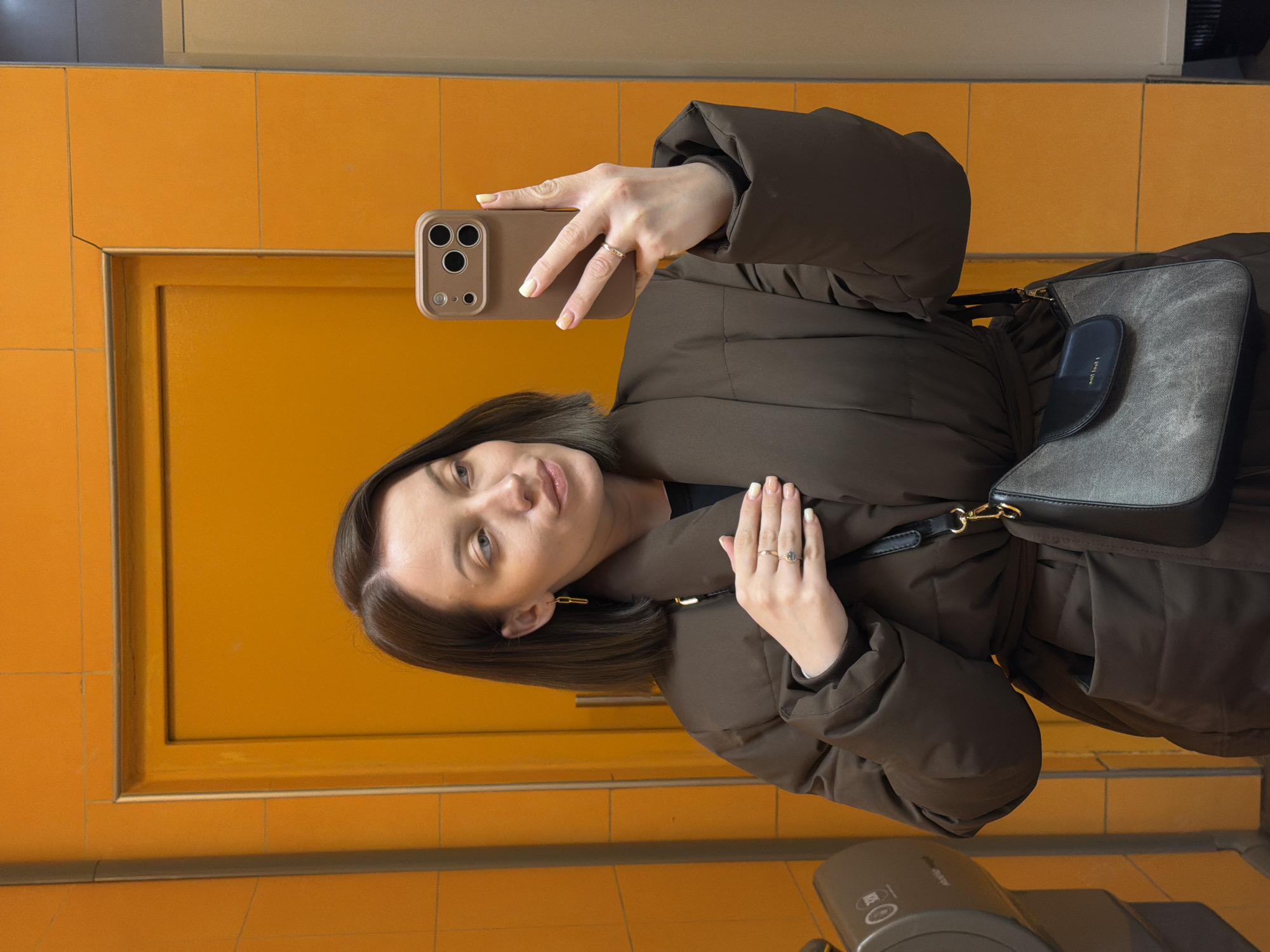 A woman with shoulder-length brunette hair is taking a selfie in a bathroom setting. She wears a brown, puffy jacket and a small grey and black purse hangs off her left shoulder. A gold ring is visible on her left hand. She holds a brown-cased iPhone in her right hand. The background consists of orange tiled walls and a door. The location in the city of Klin, Russia, cannot be definitively determined from the image. A grey object appears in the bottom of the image.