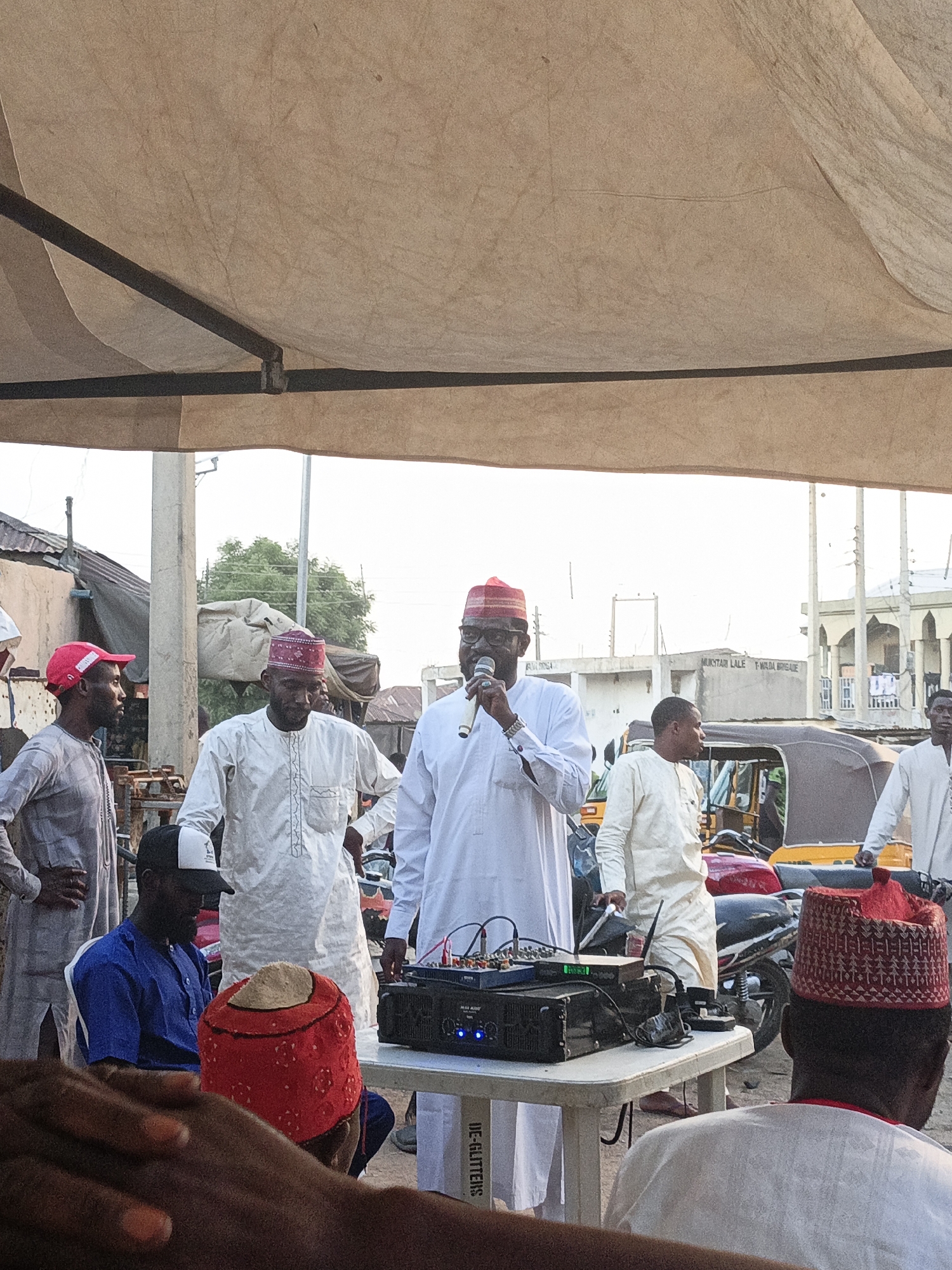 The image depicts an outdoor gathering of men under a beige canopy in Gwagwarwa, Nigeria. In the center, a man wearing a white traditional garment and a red cap, with glasses, is speaking into a silver microphone held in his right hand. To his left, another man in a white traditional garment and a red cap stands looking forward. Further left, a man in a grey traditional garment and a red baseball cap stands with his hands on his hips, looking right.
In the mid-ground, a white plastic table holds black sound equipment, including a mixer and amplifier, with various cables connected. Below the table, a white box with "WE-GLITTERS" printed vertically is visible. To the right of the table, the back of a man wearing a red patterned cap and white traditional attire is seen, with his right hand resting on the table. To the left of the table, a man wearing a blue shirt and a black and white baseball cap is seated.
In the background, several buildings of varying construction are visible under a clear sky. One building has a sign with "MUKHTAR LALE T-WADA BRIGADE" written on it. Other structures include utility poles, a mosque-like building, and various motorcycles or motorized tricycles (Keke Napep) parked. The ground appears to be unpaved or dusty. In the immediate foreground, part of a person's hands and forearm are visible at the bottom left.