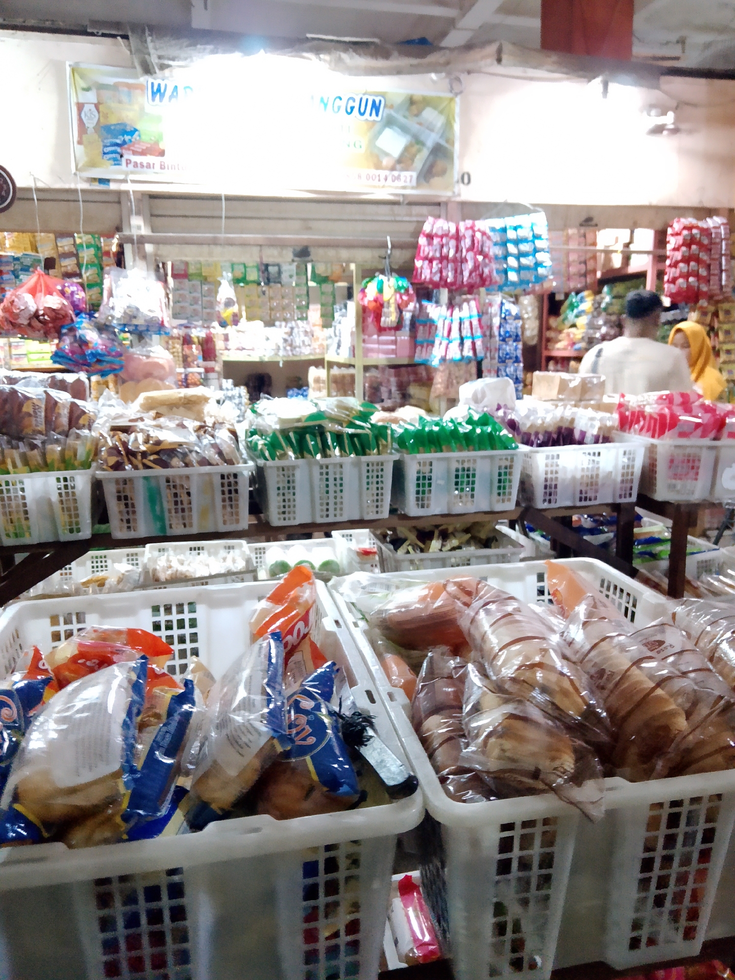 The image depicts a market stall in Tanubayan, Indonesia, likely at a Pasar Bintang. The stall is filled with various food items, predominantly packaged snacks and baked goods, displayed in white plastic baskets. In the foreground, large white baskets contain individually wrapped bread or pastry items, some with visible brown stripes, all sealed in clear plastic. To the left, another basket holds bagged snacks, with some red and blue packaging prominently visible. In the mid-ground, several smaller white baskets are neatly arranged on shelves. These baskets contain an assortment of packaged items, including green-packaged snacks, a basket of purple-packaged items, and another filled with red-packaged goods. Further back, more shelves are stocked with various packaged products. The stall has a banner at the top with Indonesian text. Two individuals are partially visible in the background; one appears to be a person in a light-colored shirt, and another, a person wearing a yellow headscarf, is standing behind them. The overall environment suggests a bustling, well-stocked market.