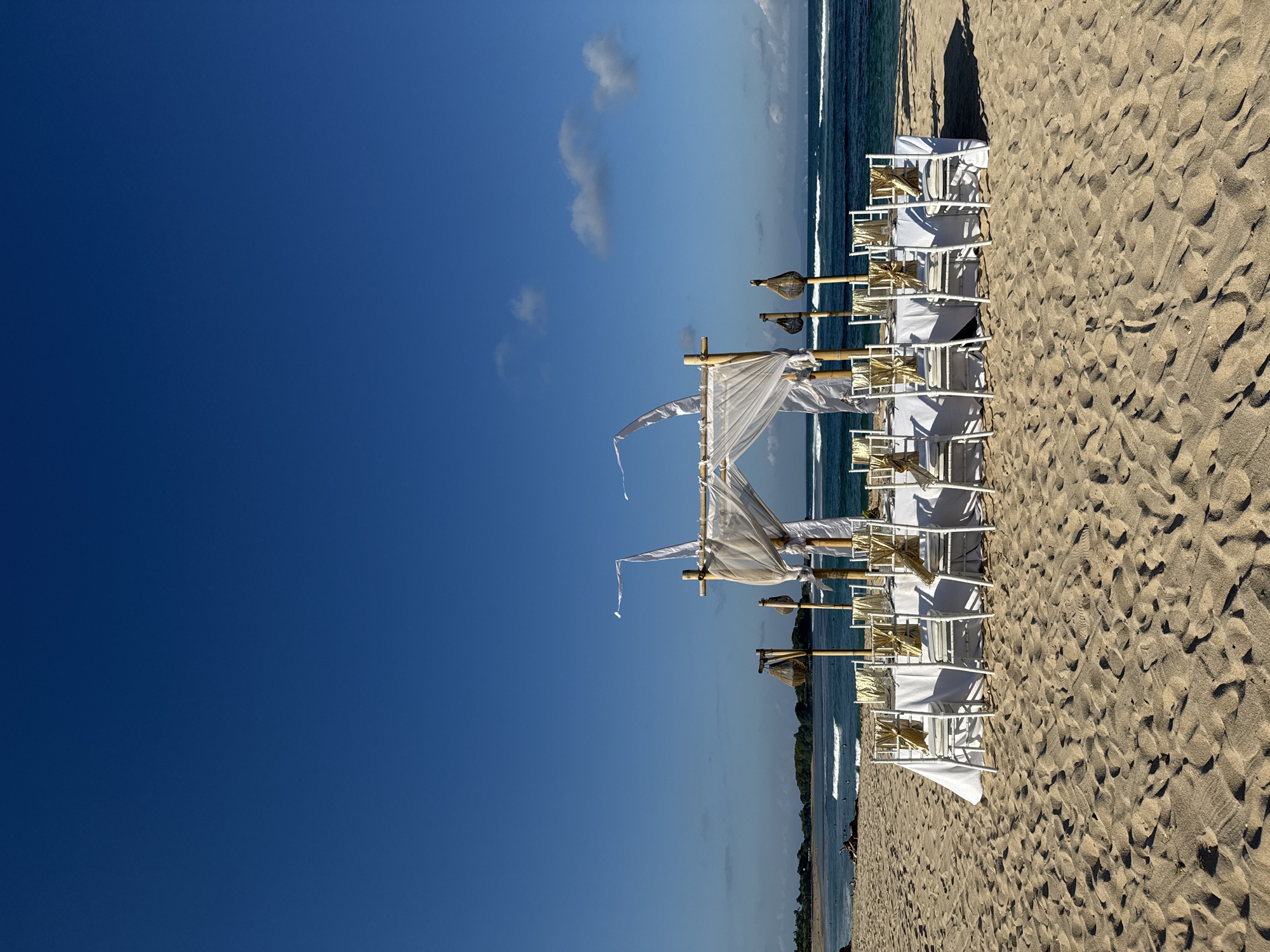 This image captures a serene beach setting in Kuta Selatan, Indonesia, likely prepared for a special event such as a wedding or ceremony. The photograph is oriented vertically, but should be viewed as a horizontal landscape with a vast blue sky at the top, and the ocean and beach occupying the lower portion of the frame.
On the right side of the sandy beach, a meticulously arranged setup faces the calm, blue-green ocean. A central archway, constructed from natural bamboo poles, is elegantly draped with sheer white fabric that billows gently, suggesting a light breeze. Leading up to this arch, multiple rows of white chairs are neatly aligned, each adorned with a light-colored (possibly golden or beige) sash on its backrest. Interspersed along the aisle and near the seating are tall, rustic poles topped with basket-like structures, possibly lanterns or torches, adding to the decorative ambiance.
The sand exhibits clear textures and shadows, indicating bright daytime lighting, likely morning or late afternoon, given the length and angle of the shadows cast by the chairs and structures. The sky above is a brilliant, clear blue with a few wispy white clouds. The ocean surface shows gentle waves breaking in the distance, and a faint landmass or coastline is visible along the horizon on the far left, suggesting a bay or curved shoreline. No people or visible text are present, allowing the natural beauty and elegant preparation to dominate the scene, evoking a sense of peaceful anticipation.