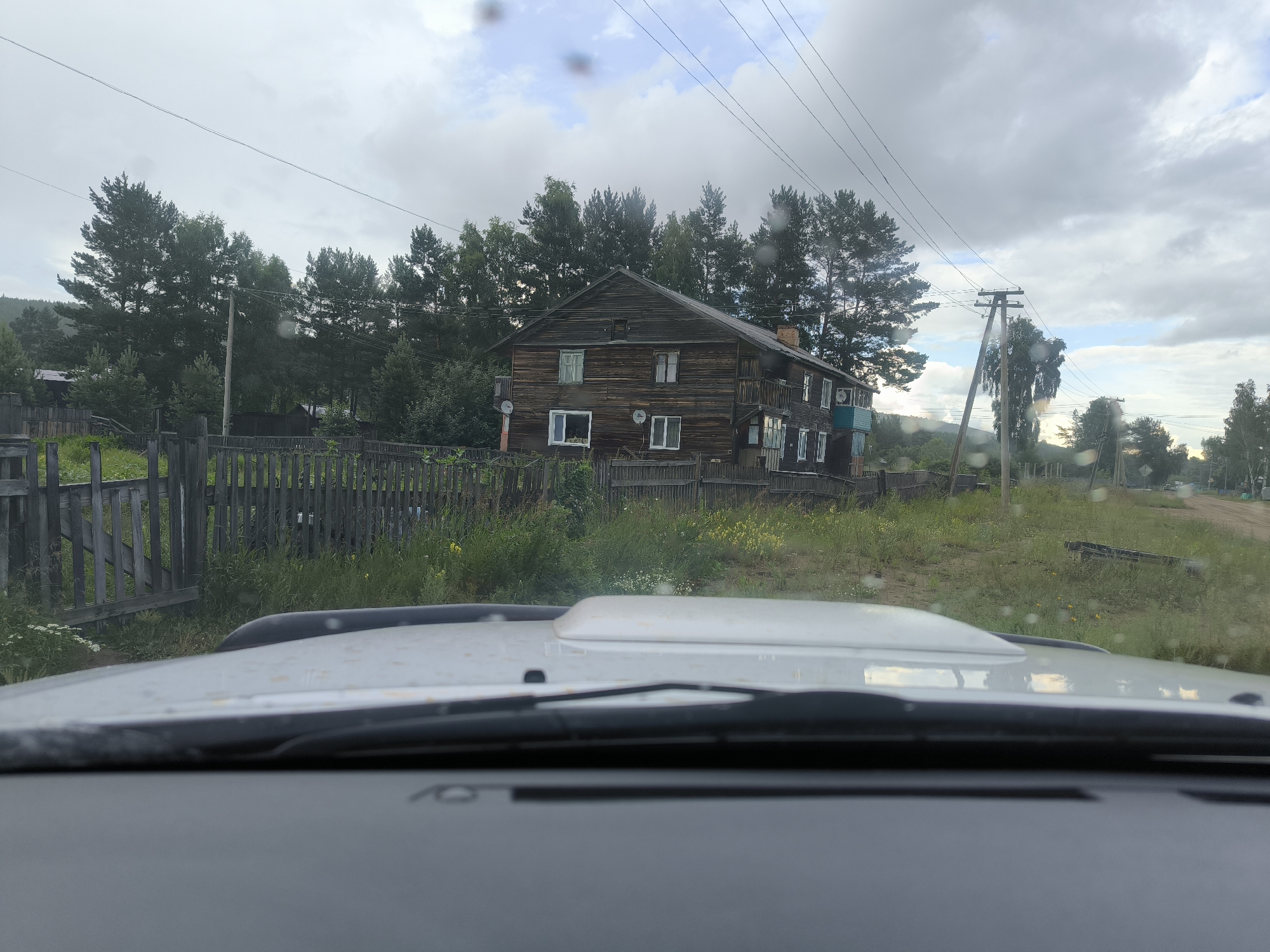 The image shows a view from inside a vehicle, looking out at a wooden house in the city of Gremiuchiy, Russia. The house is two stories tall with a dark wooden exterior and multiple windows. A wooden fence runs in front of the house, and there are trees and a dirt road in the background. The sky is cloudy.