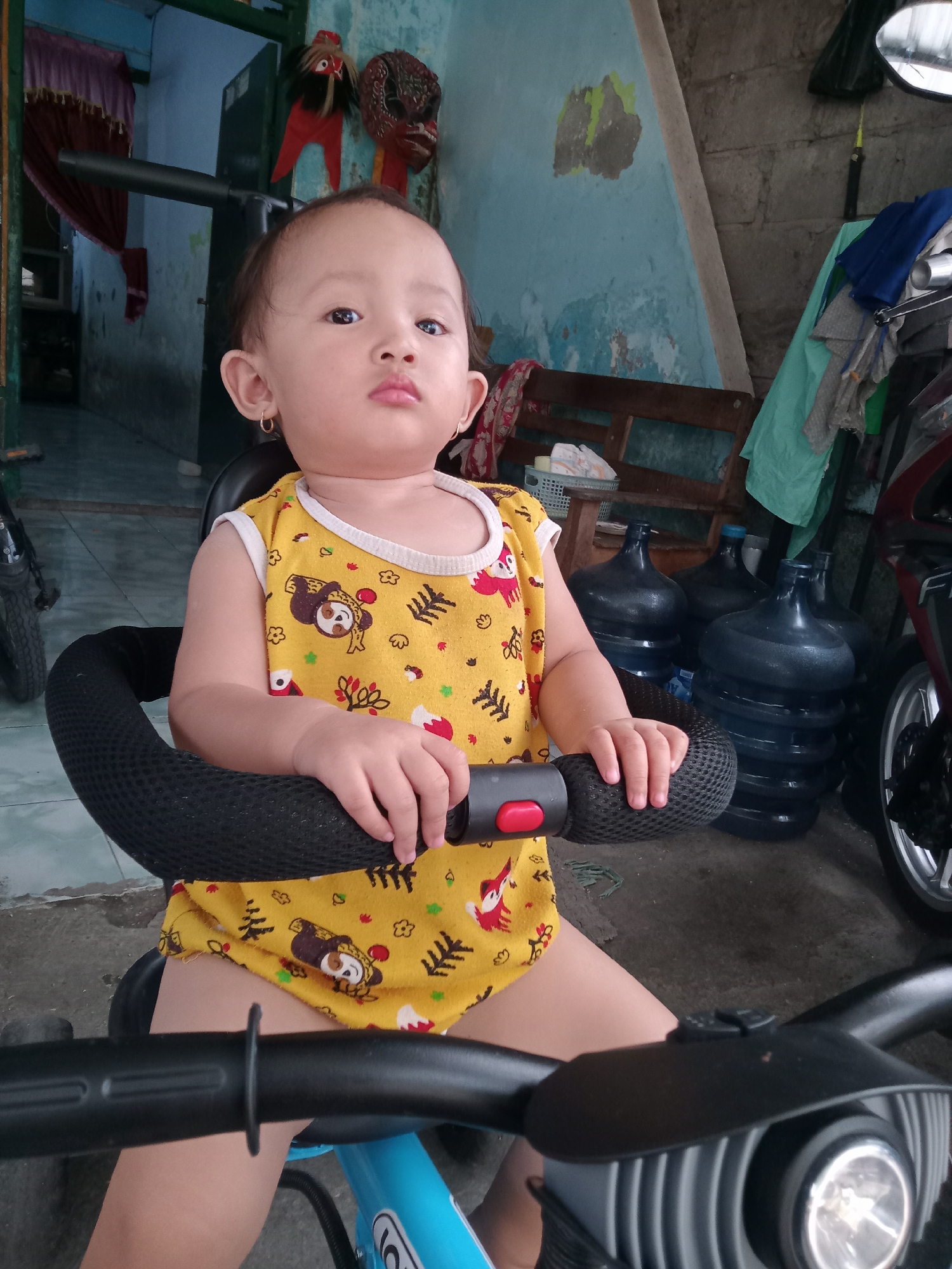 A young child, likely a toddler, is the central subject of the image, sitting on a blue and black ride-on toy or tricycle. The child wears a yellow sleeveless top with cartoon animal patterns, and small gold hoop earrings are visible. They look directly at the camera with a neutral, slightly pensive expression, holding onto the black handlebars of the toy.
The setting appears to be a sheltered outdoor or indoor area, possibly a porch or garage, given the varied background elements. To the left, a dark-colored tire from another vehicle is partially visible. Further back, two traditional Indonesian masks, one red and black and another brown, hang on a light blue wall which shows signs of wear with some paint peeling. A dark red curtain hangs to the left of the masks, partially obscuring a doorway or opening.
To the right of the child, several large, dark blue plastic water dispenser bottles are stacked on the floor. Beyond them, parts of a red and black motorcycle or scooter are visible, including a wheel and fender. A wooden bench or shelf is also seen in the background to the right. The floor beneath the child and bottles is a light-colored tiled surface. The overall environment suggests a domestic, lived-in space. There is no discernible text visible in the image.